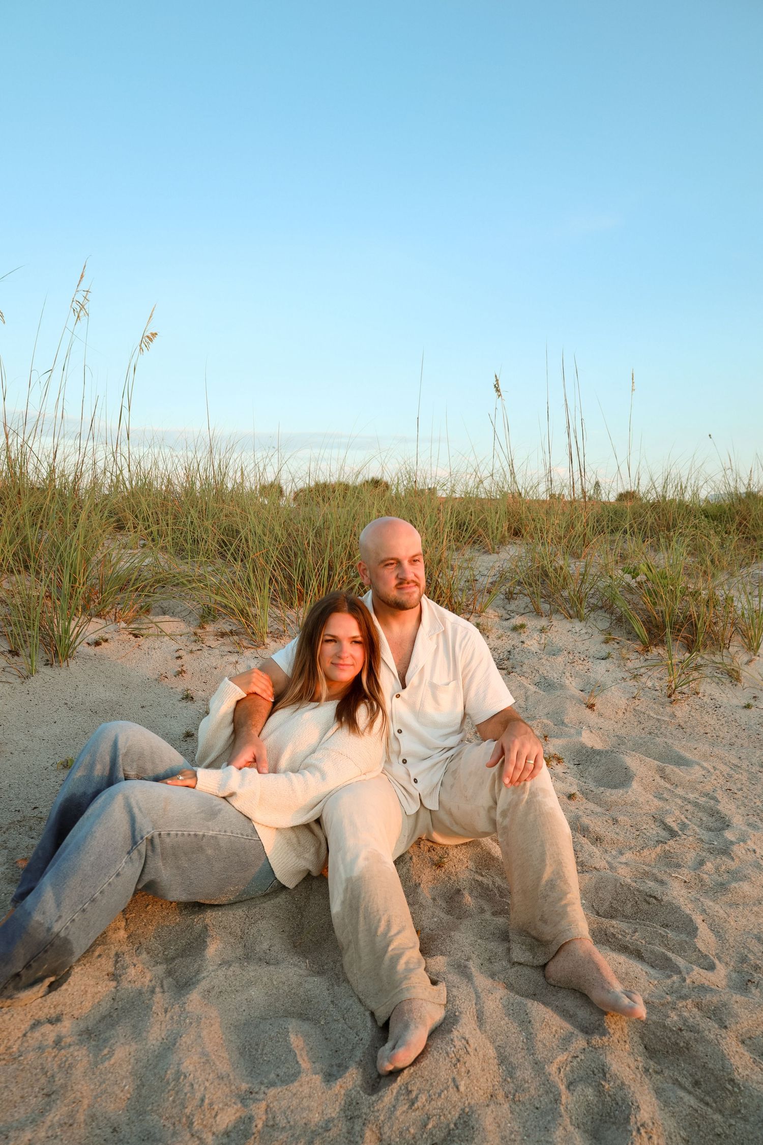 A couple sits together on sandy beach dunes at golden hour, dressed in matching linen outfits, with sea grass in the background.