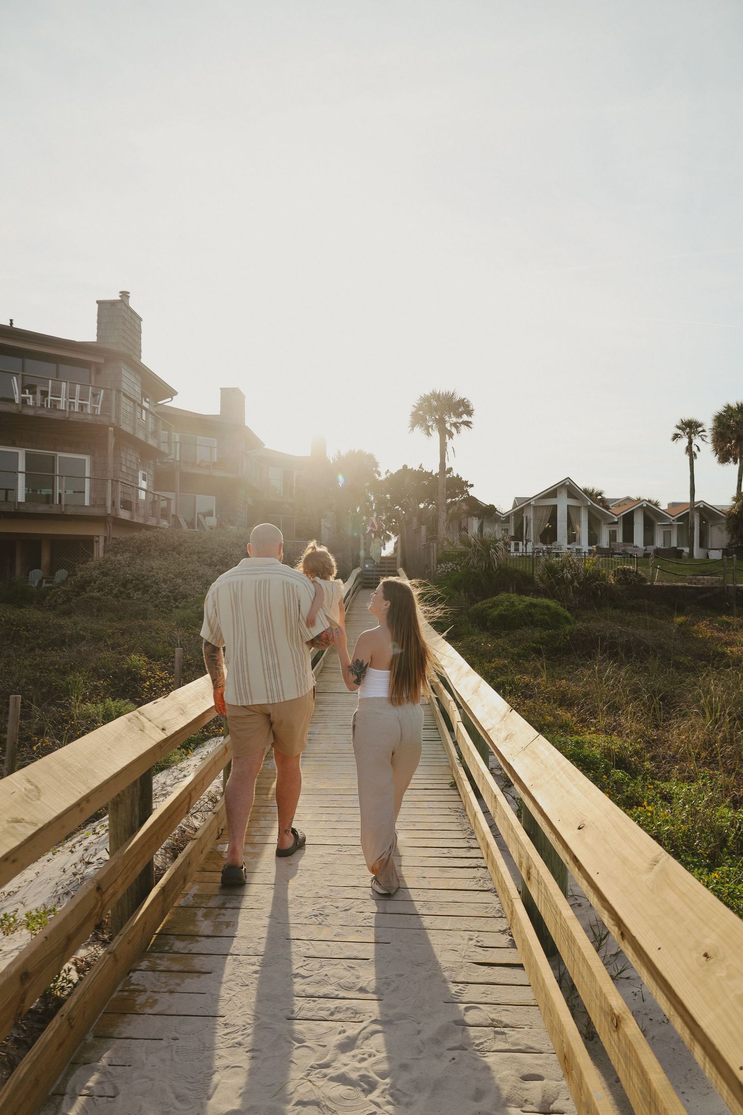 Couple walking on wooden boardwalk at sunset with beach houses in background.