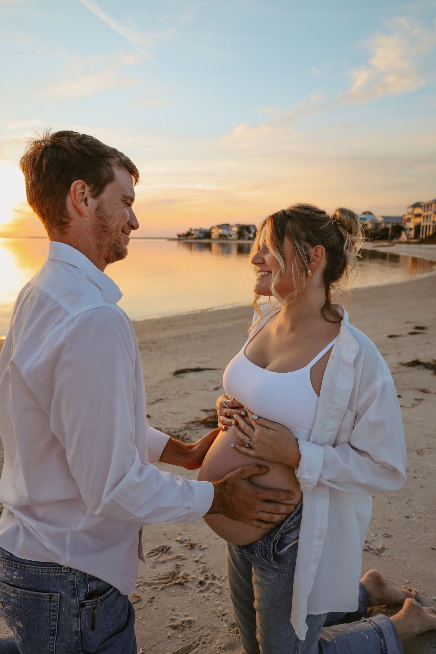 Romantic couple embraces on a sandy beach at sunset with waves and beach houses in the background.