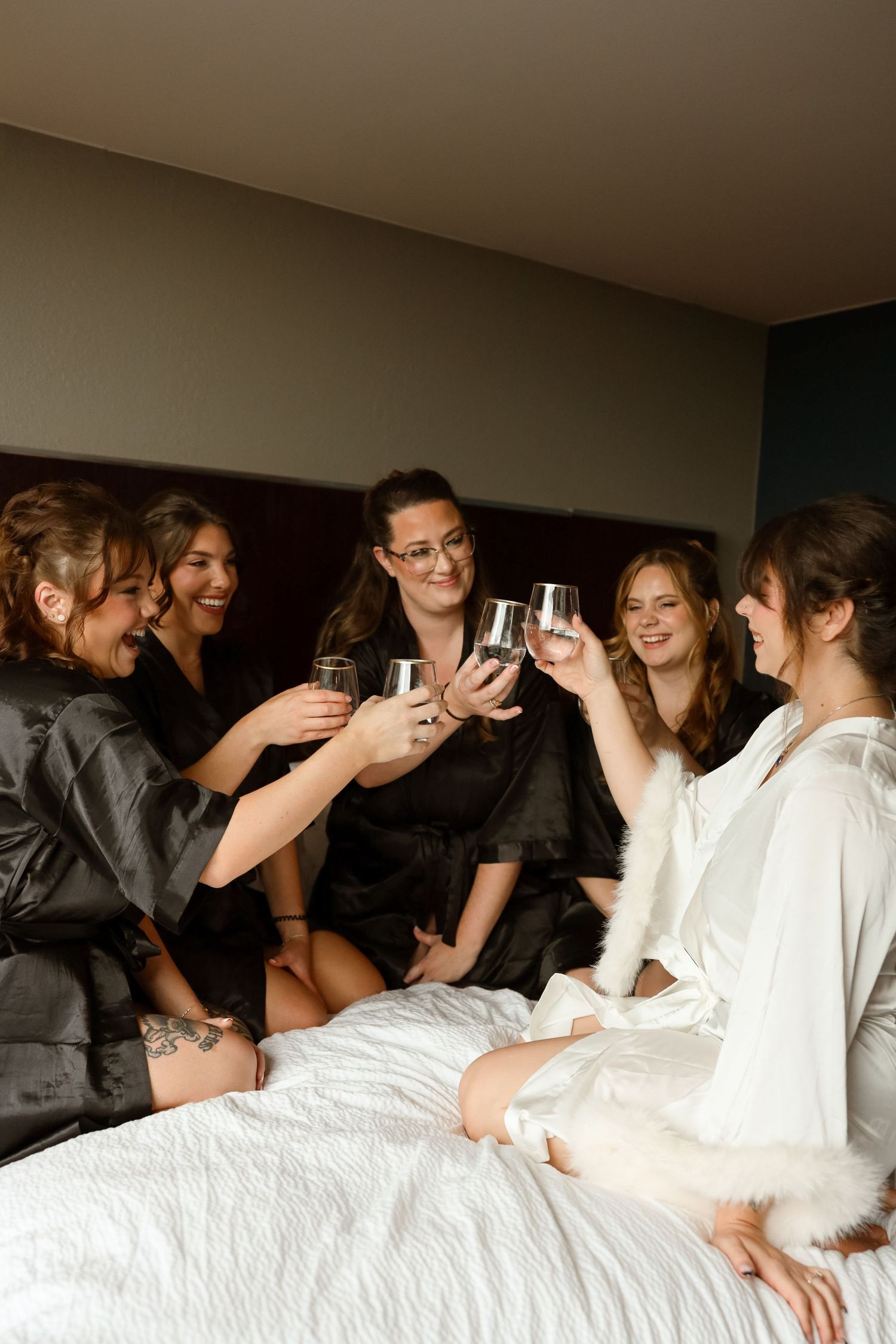 Bridesmaids in satin robes toasting with wine glasses on a bed during bridal party getting ready celebration.