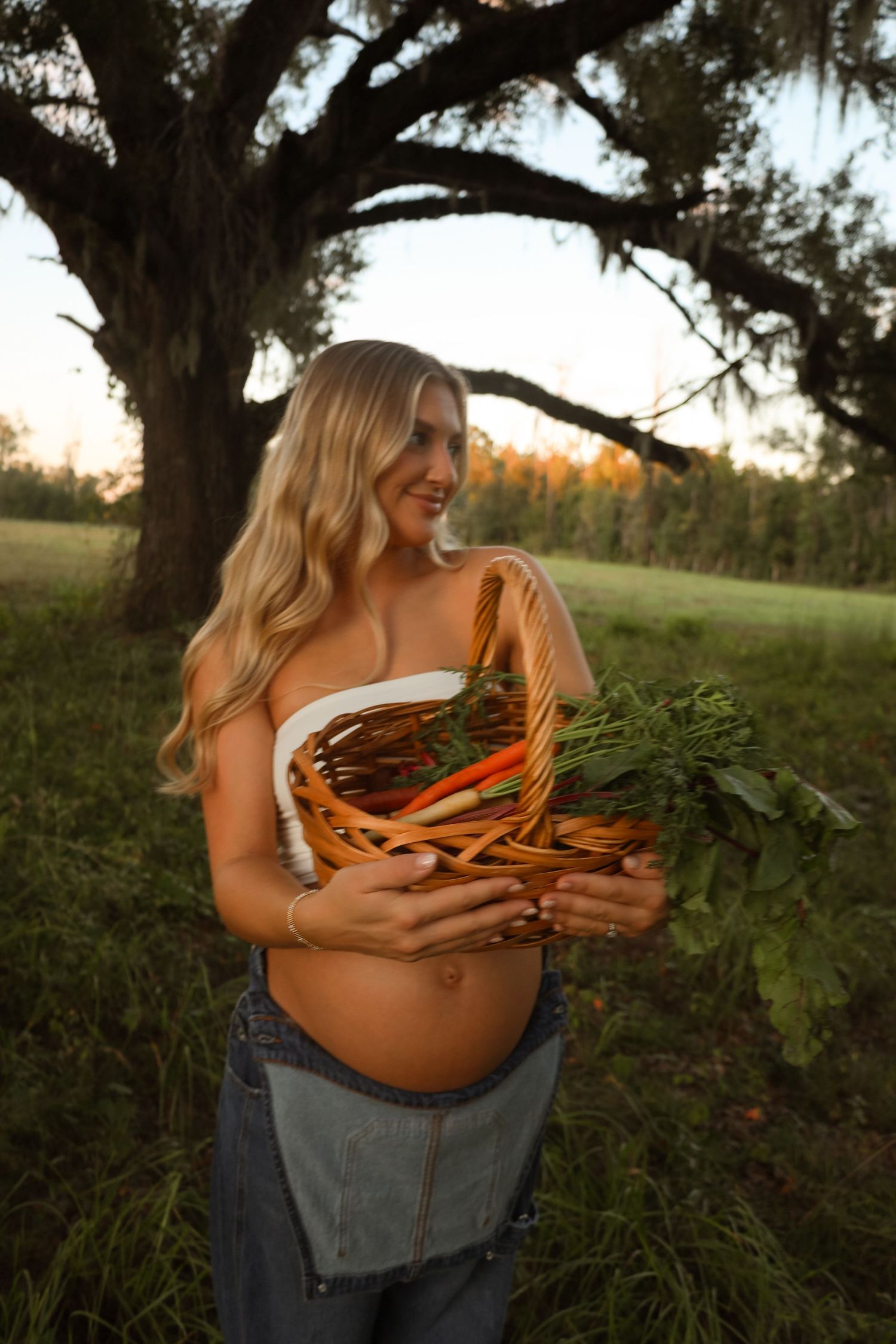 Smiling woman with long blonde hair holds a basket of fresh carrots and greens beneath a large oak tree in a grassy field at golden hour.
