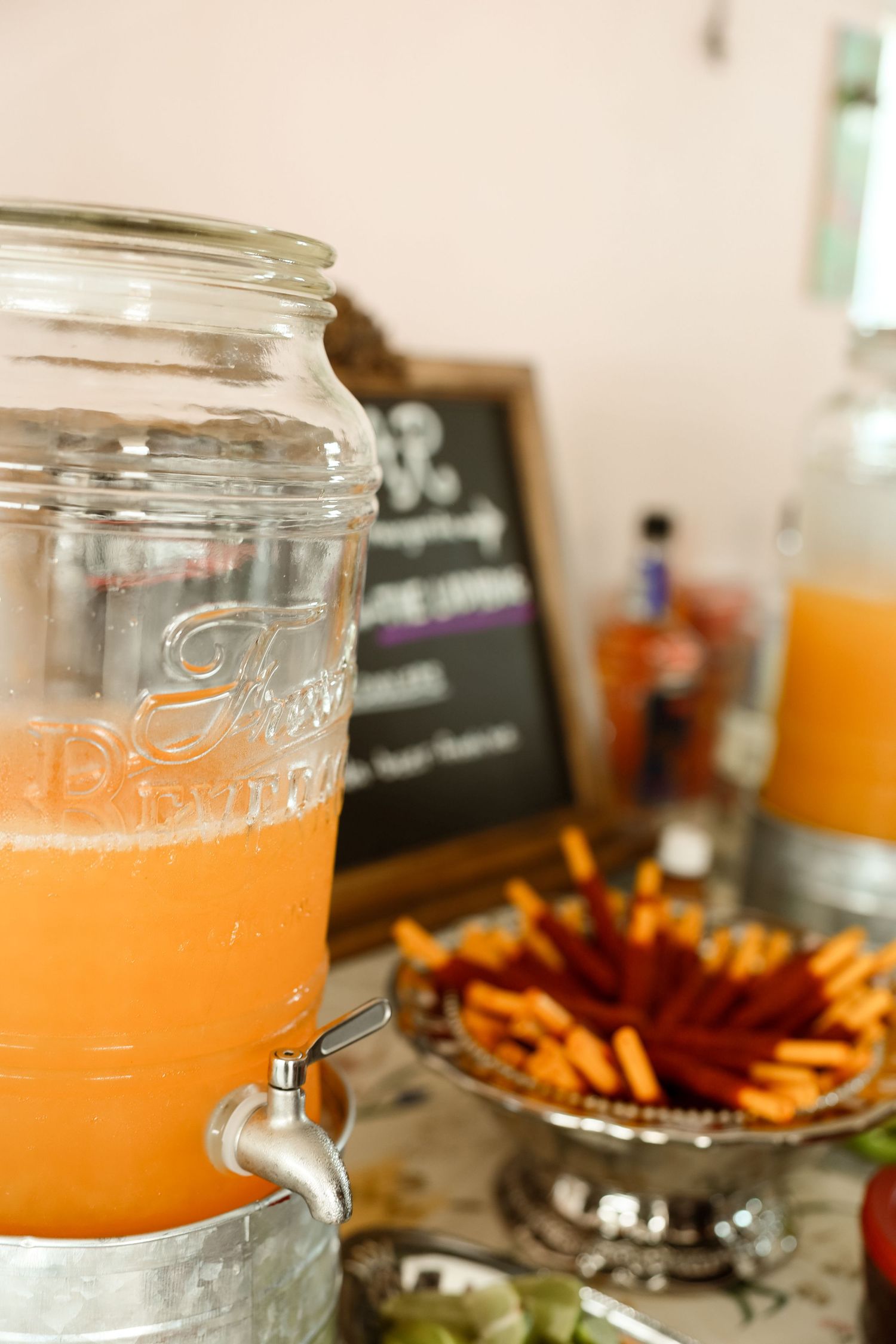 Glass drink dispenser filled with orange punch beside a silver tray of pretzel sticks at a party buffet table.