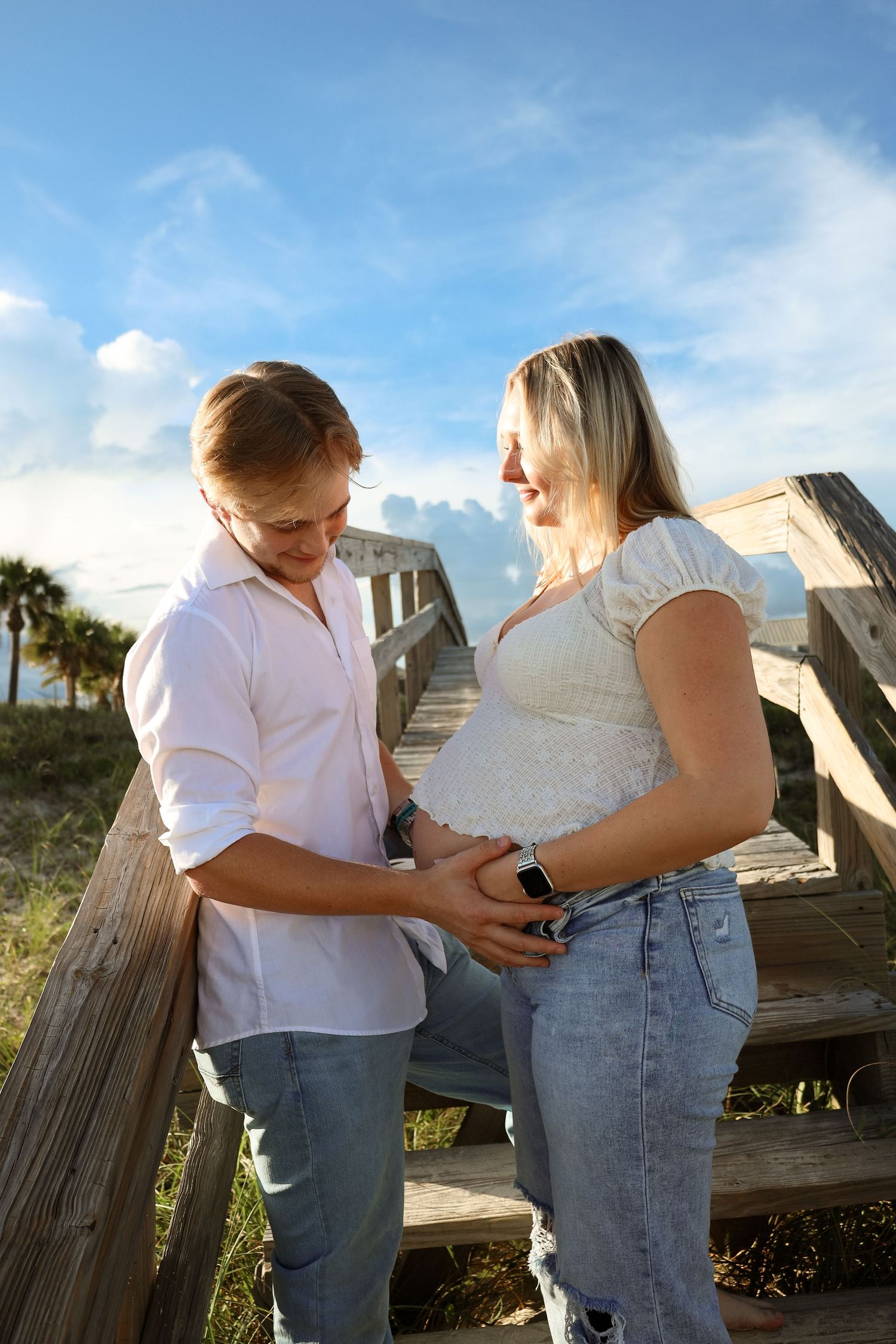 Couple at beach boardwalk, man tenderly touching pregnant woman's belly, both smiling, wearing white tops and jeans.