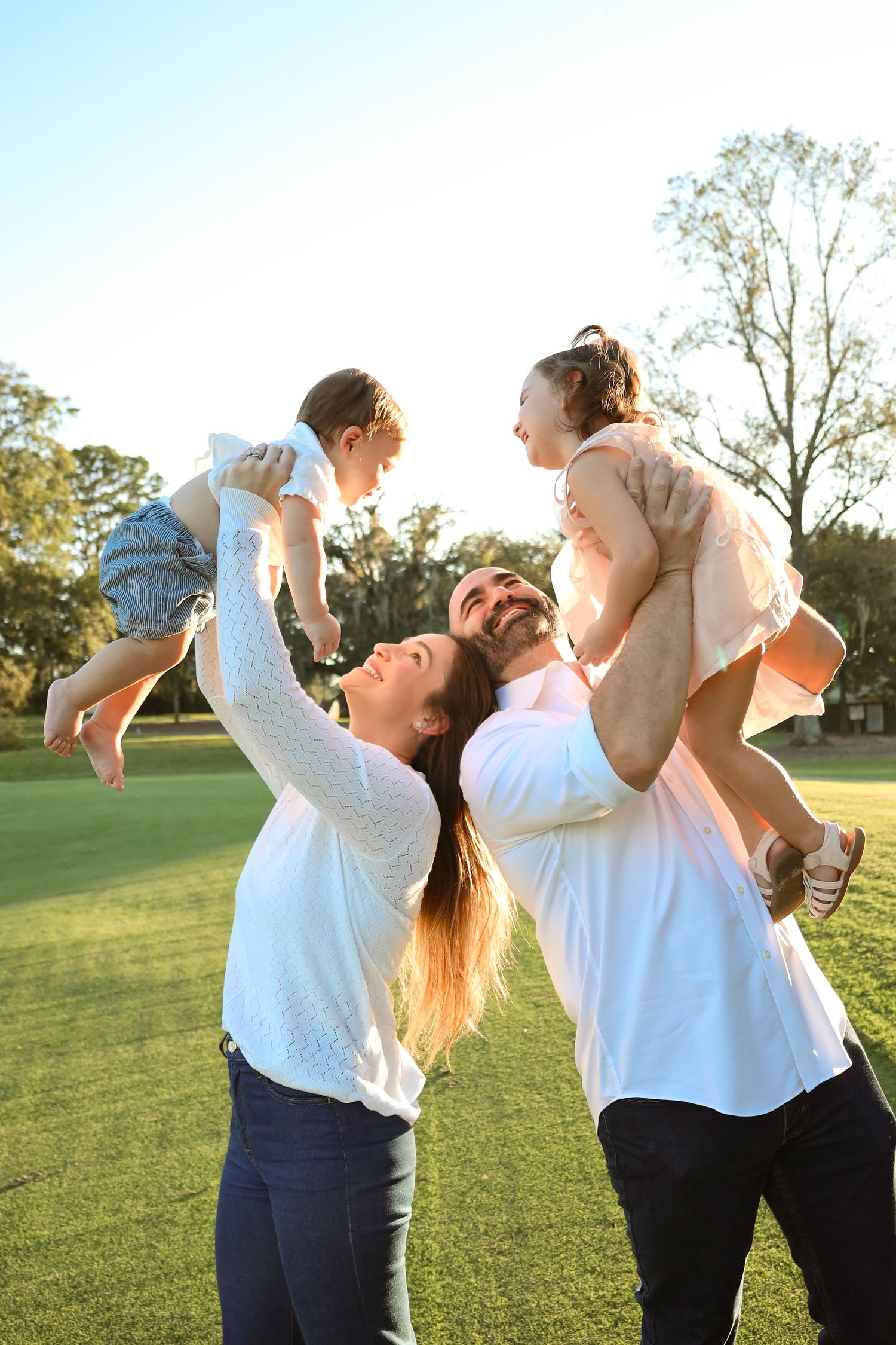 A smiling mother and father lift their two laughing young children into the air on a sunny, tree-lined grassy lawn.