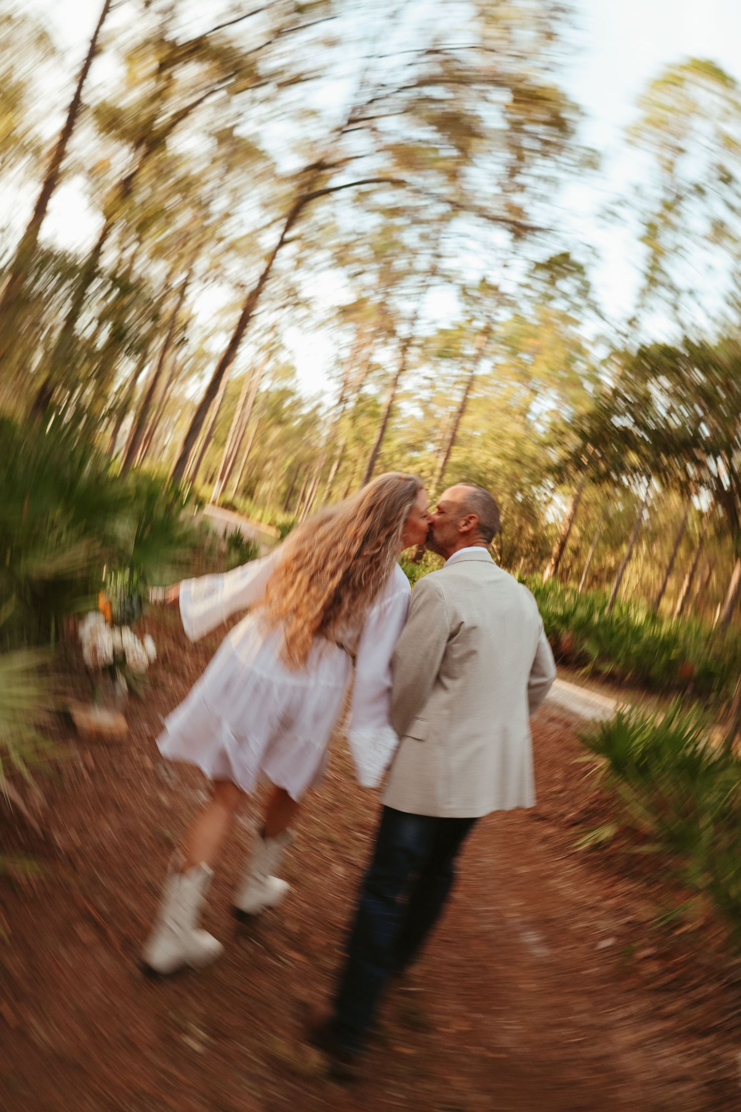 A couple in wedding attire spins and dances together on a dirt path surrounded by blurred trees at sunset.