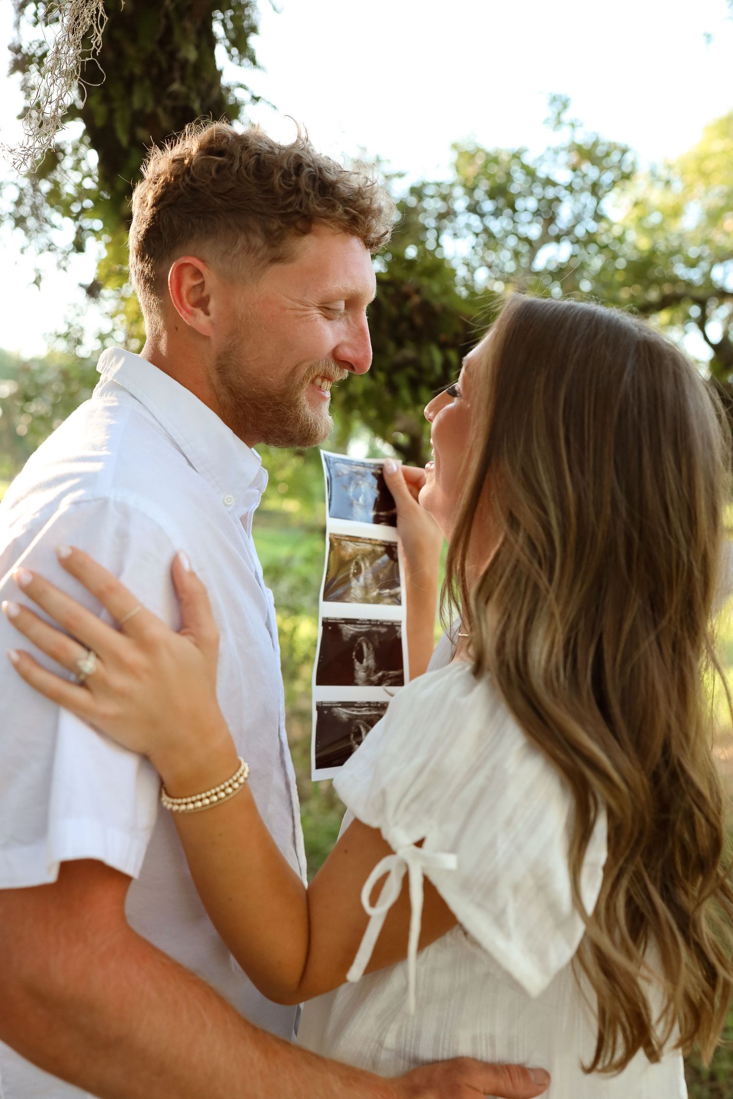 Happy couple embracing outdoors holding ultrasound photos, announcing pregnancy in a sunny garden setting.