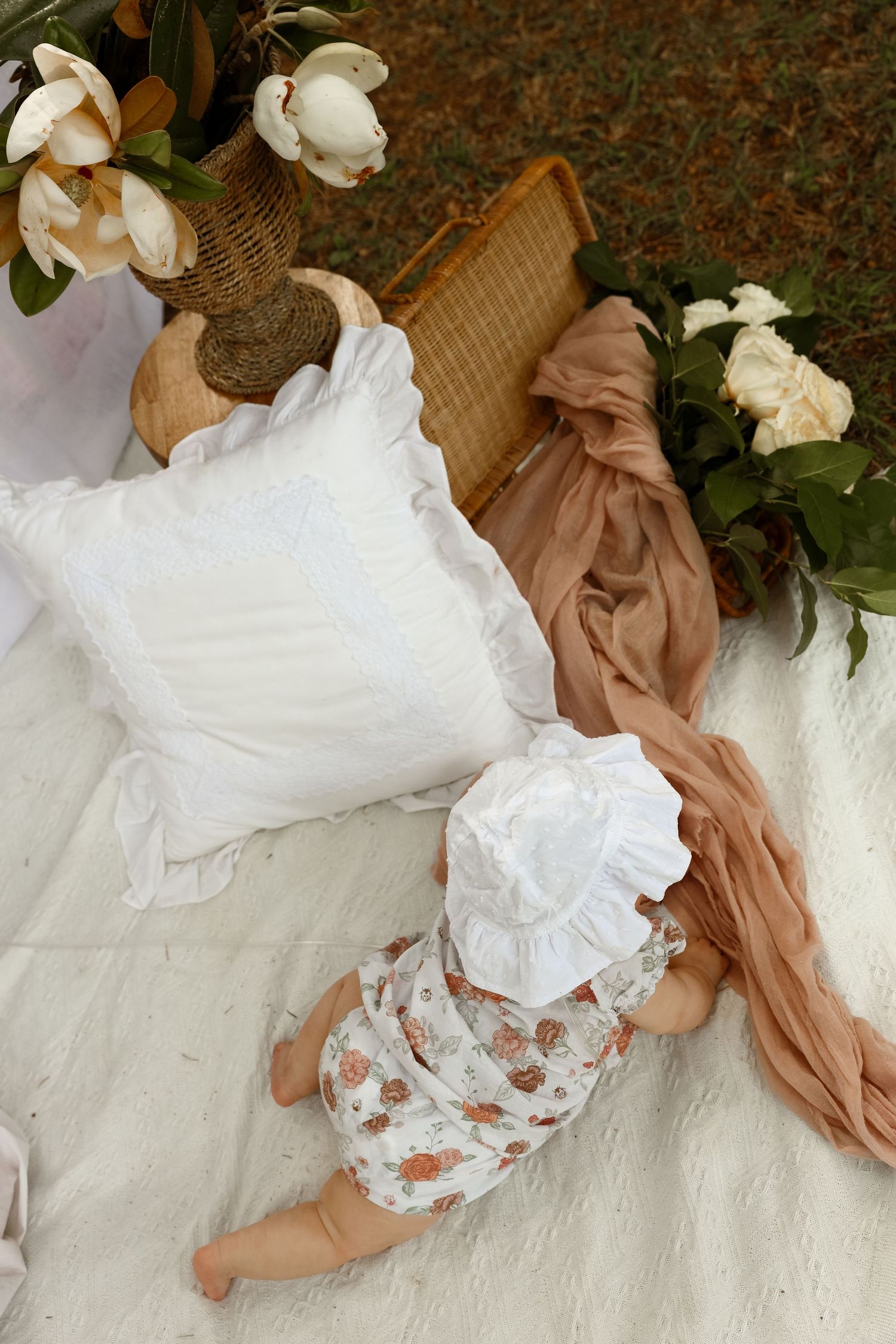 Baby in floral romper and white bonnet lying on picnic blanket with white pillow and roses.