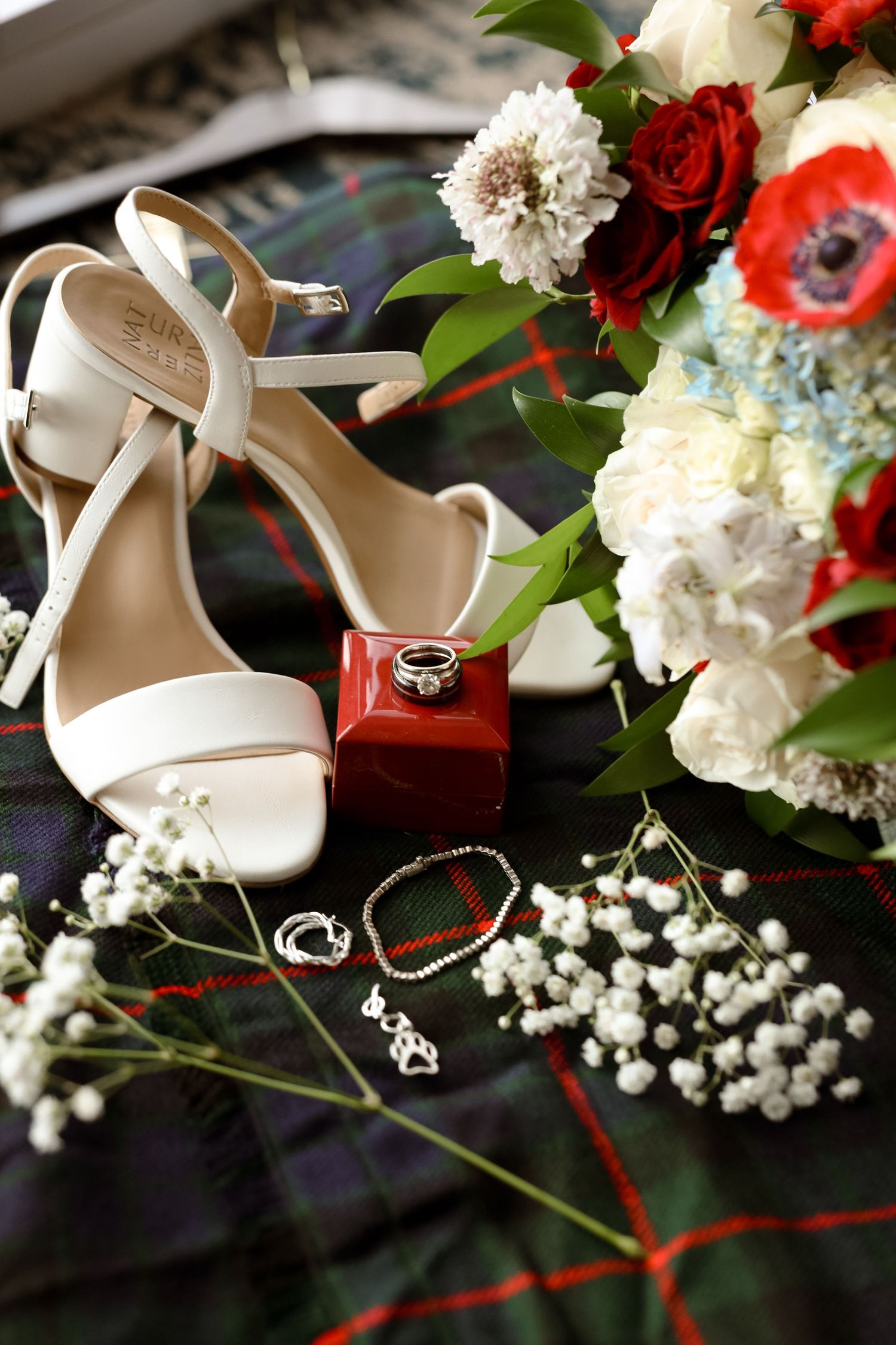 White bridal heels, diamond ring on red box, pearl bracelet, and red and white floral bouquet on plaid fabric.