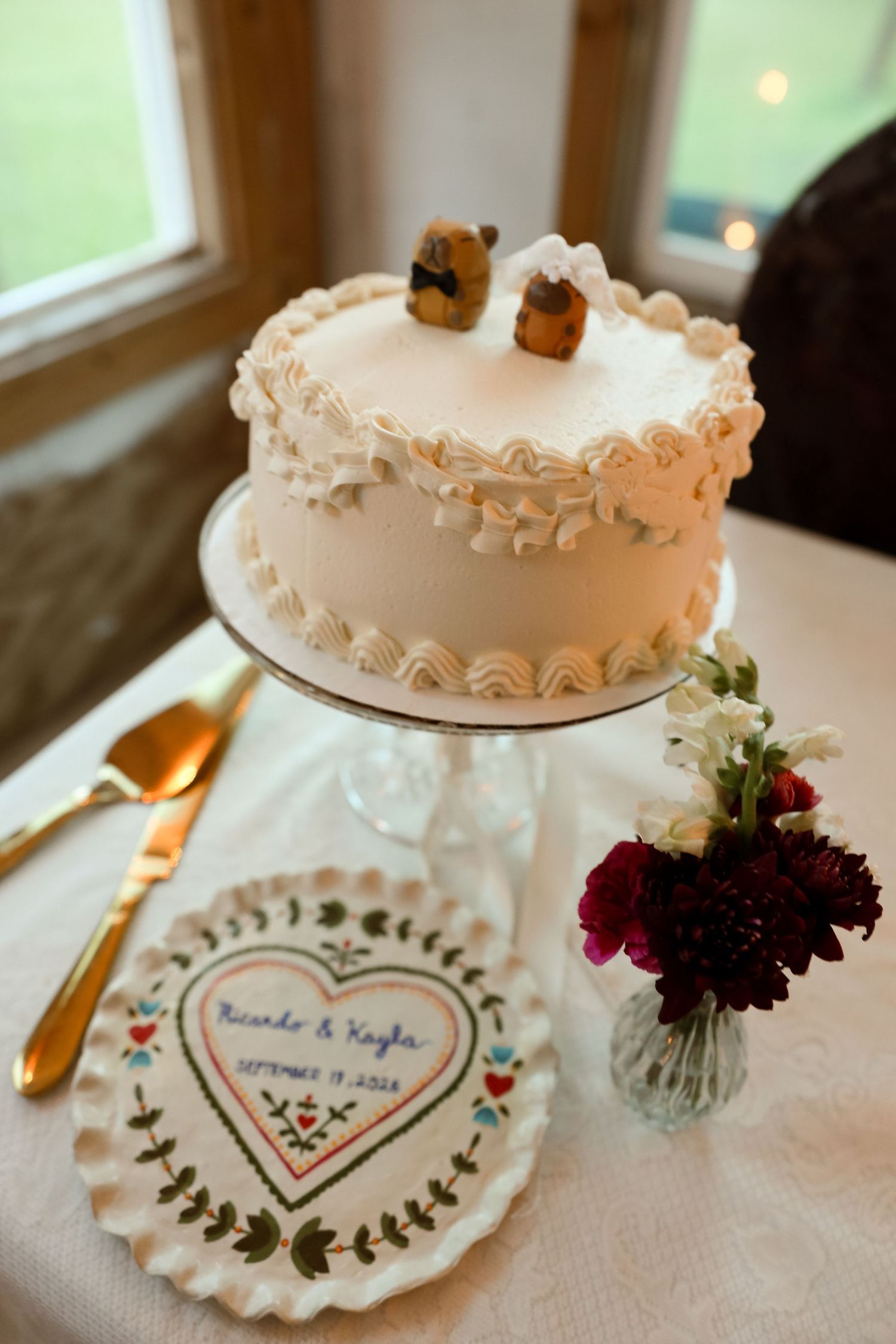 Elegant white wedding cake with bear toppers, gold server, floral arrangement, and personalized heart embroidery on display.