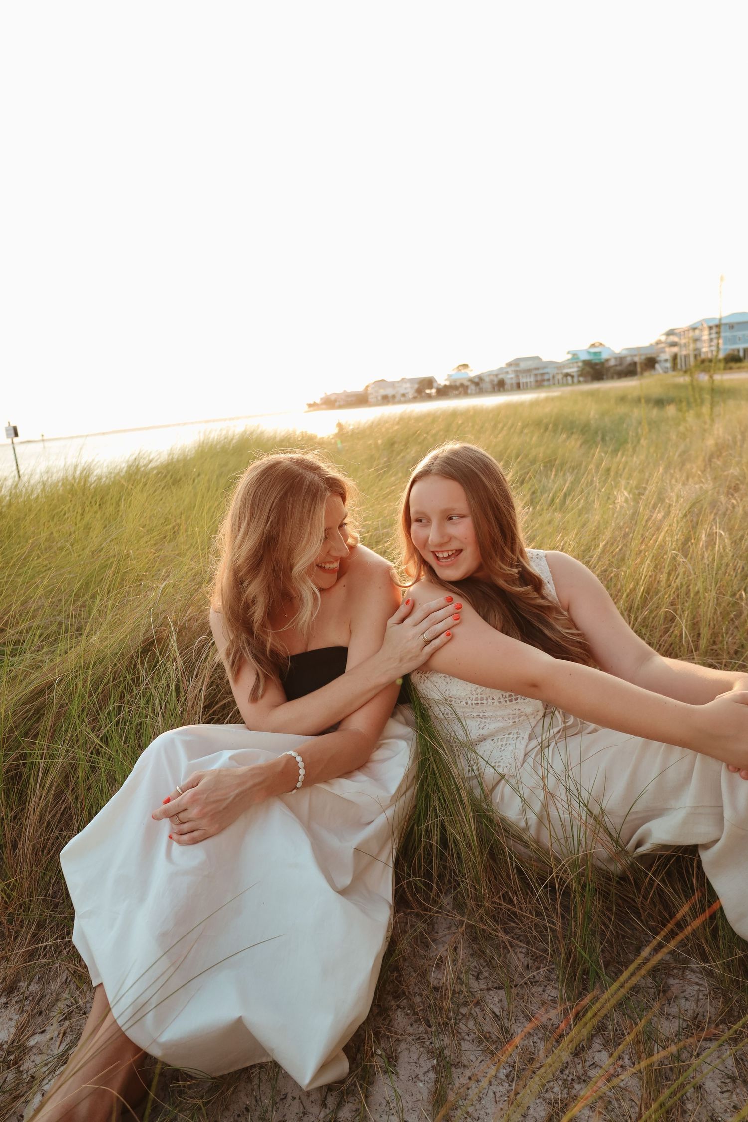 Two people in white dresses share intimate moments while sitting in tall grass at sunset.