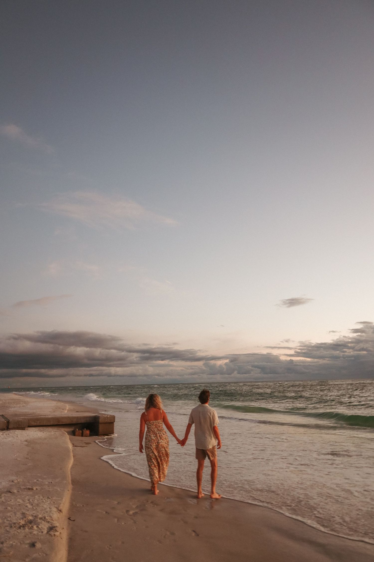 A couple walks hand in hand along a serene beach shoreline during a cloudy sunset.