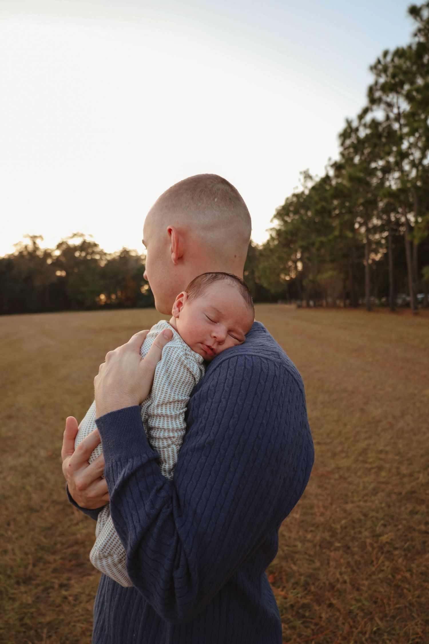 Parent in navy sweater holds baby in striped outfit against autumn field at sunset.