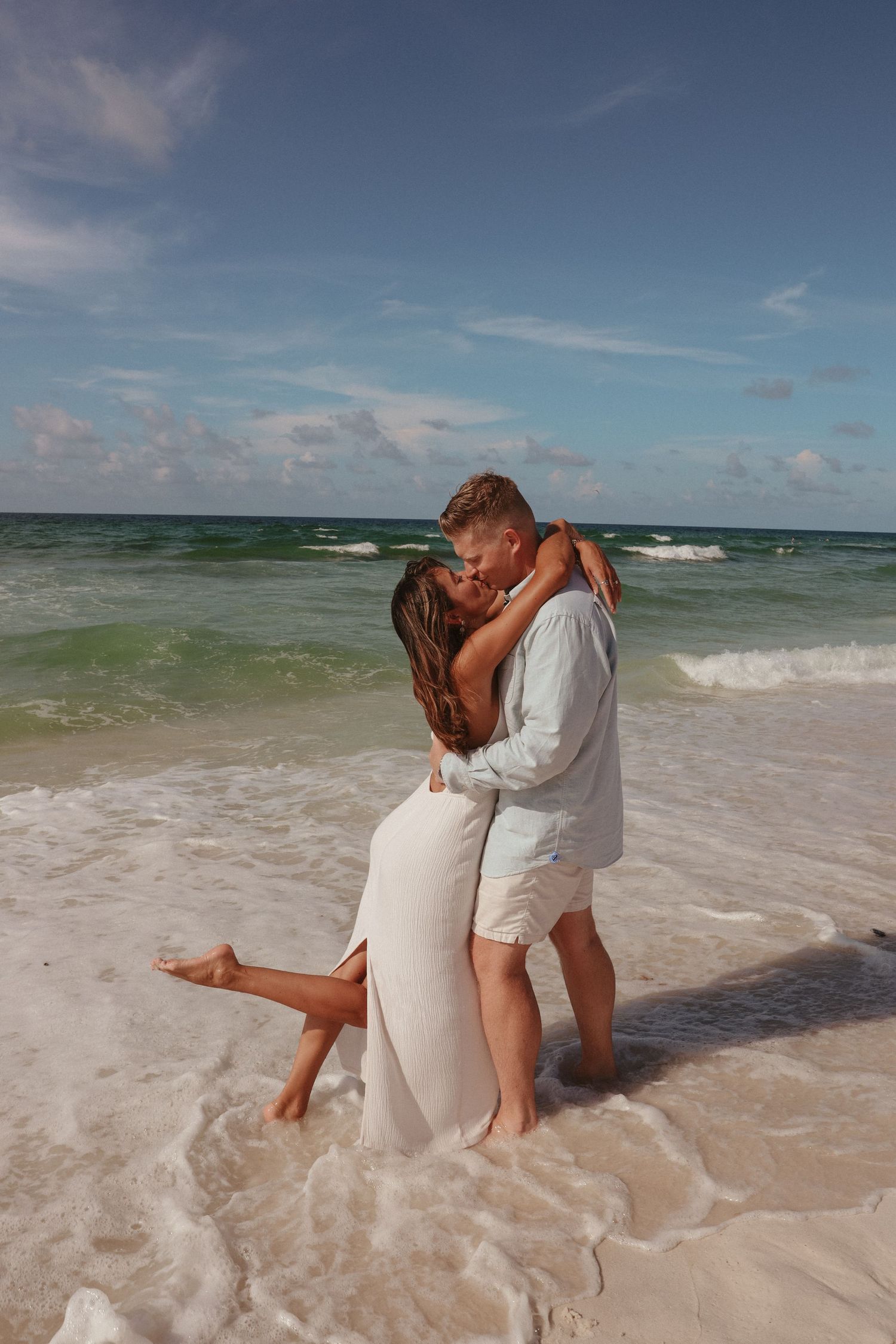 Romantic couple embraces on a sandy beach with turquoise waves and blue sky in the background.
