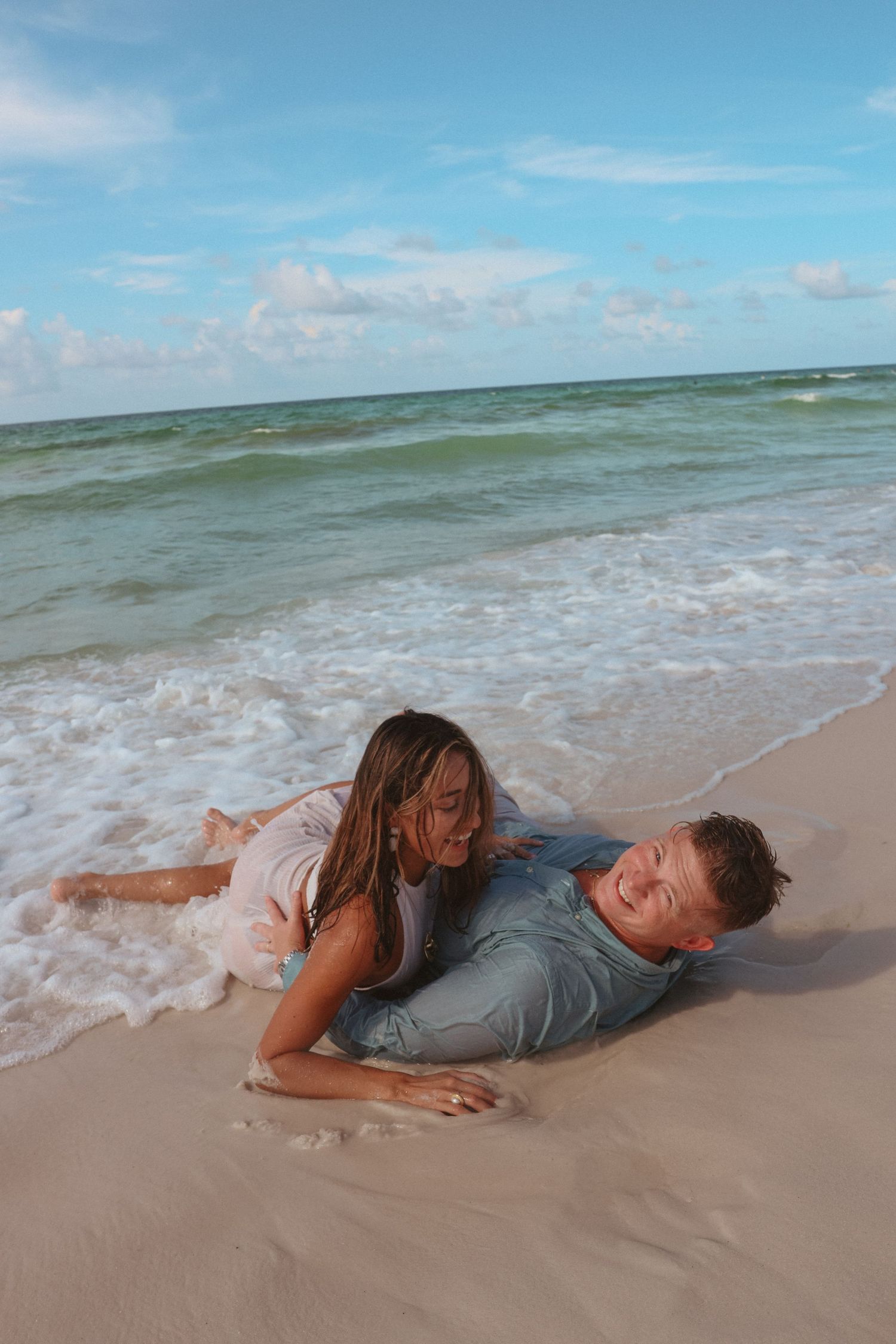 A playful couple lying on the sandy beach as waves roll in behind them.