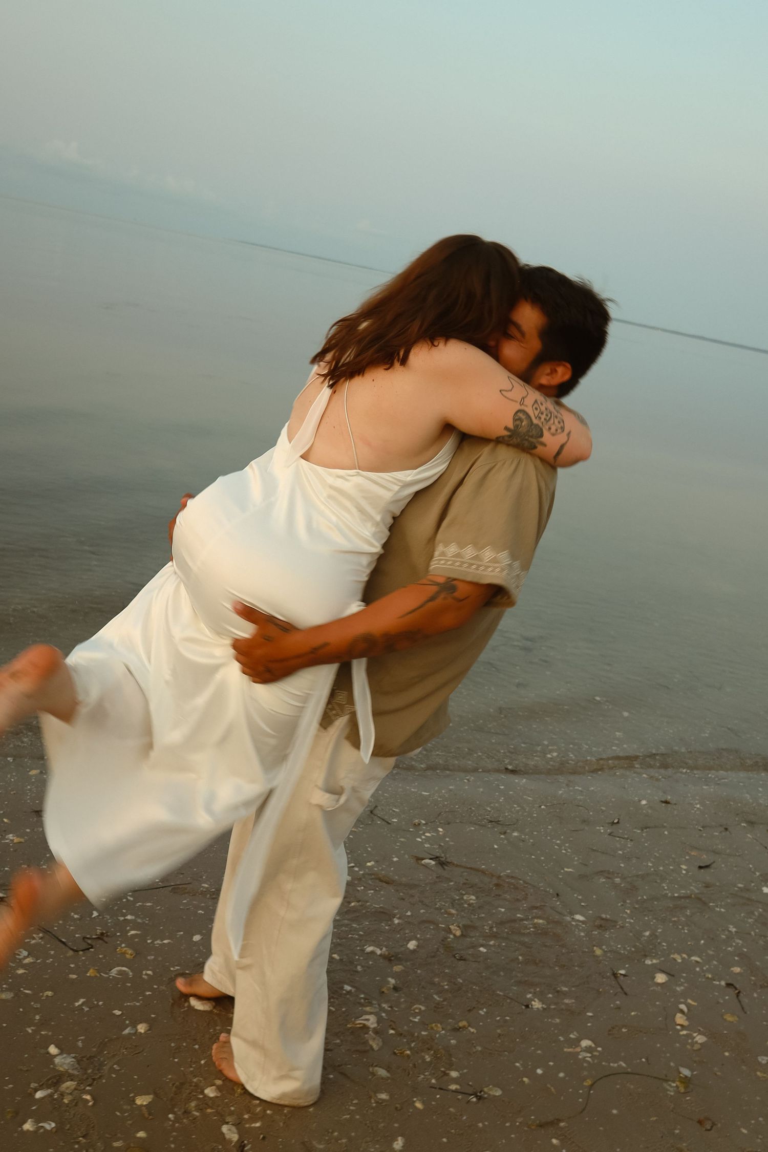 Couple embracing romantically on a beach at dusk, woman in white dress lifted by man with tattoo near calm water.