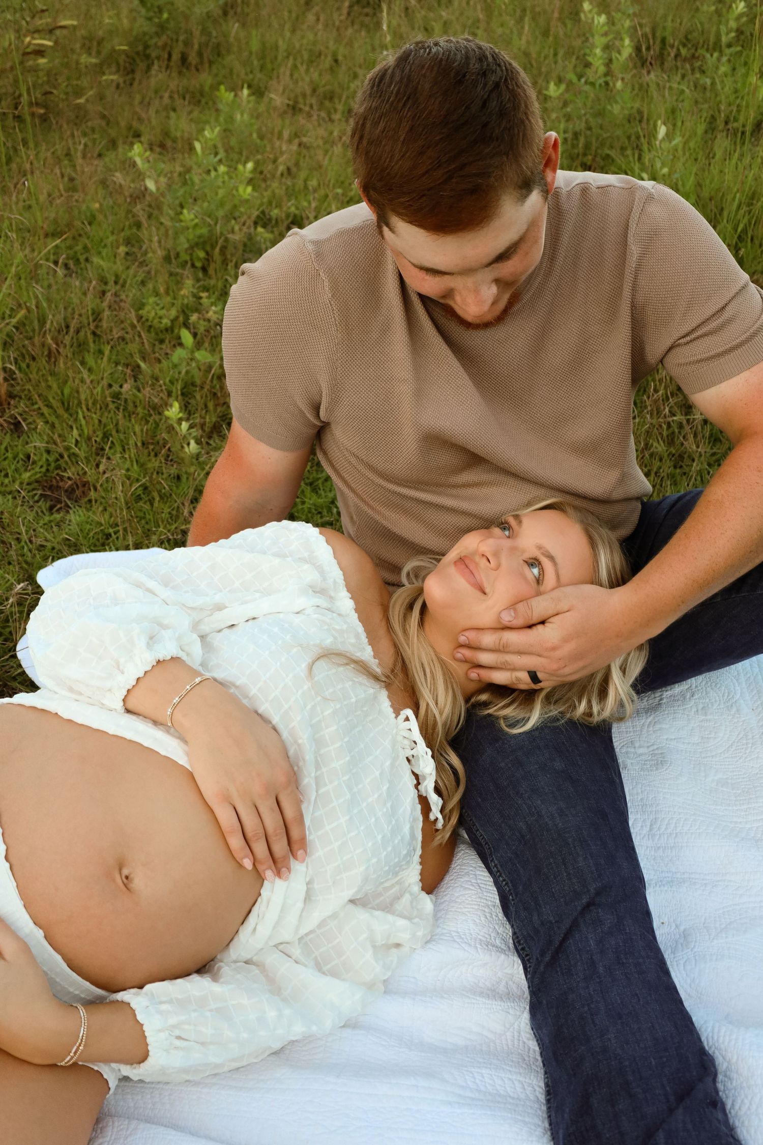 Pregnant woman in a white lace top rests her head on her partner's lap on a blanket in a grassy field during a maternity photoshoot.