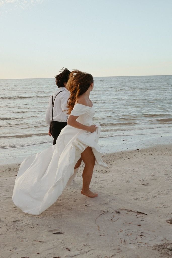 A couple embraces playfully on a sandy beach at sunset while wearing a flowing white dress and casual attire.