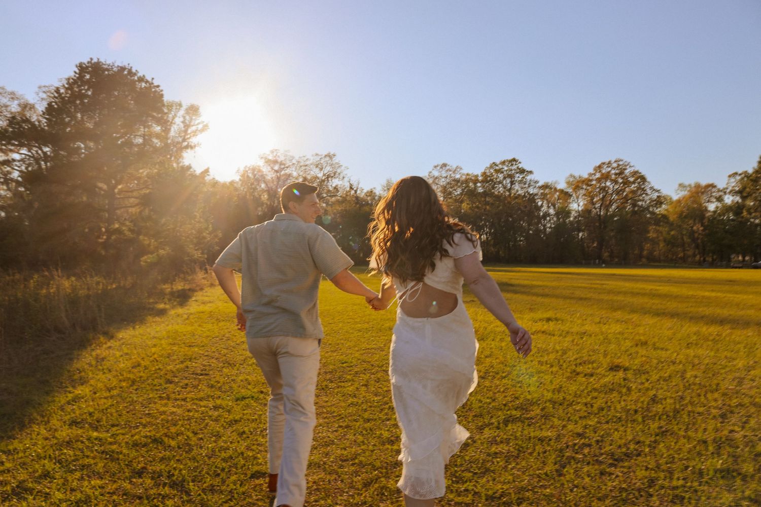 Two people in white clothing walk together across a sunlit grassy field at golden hour.