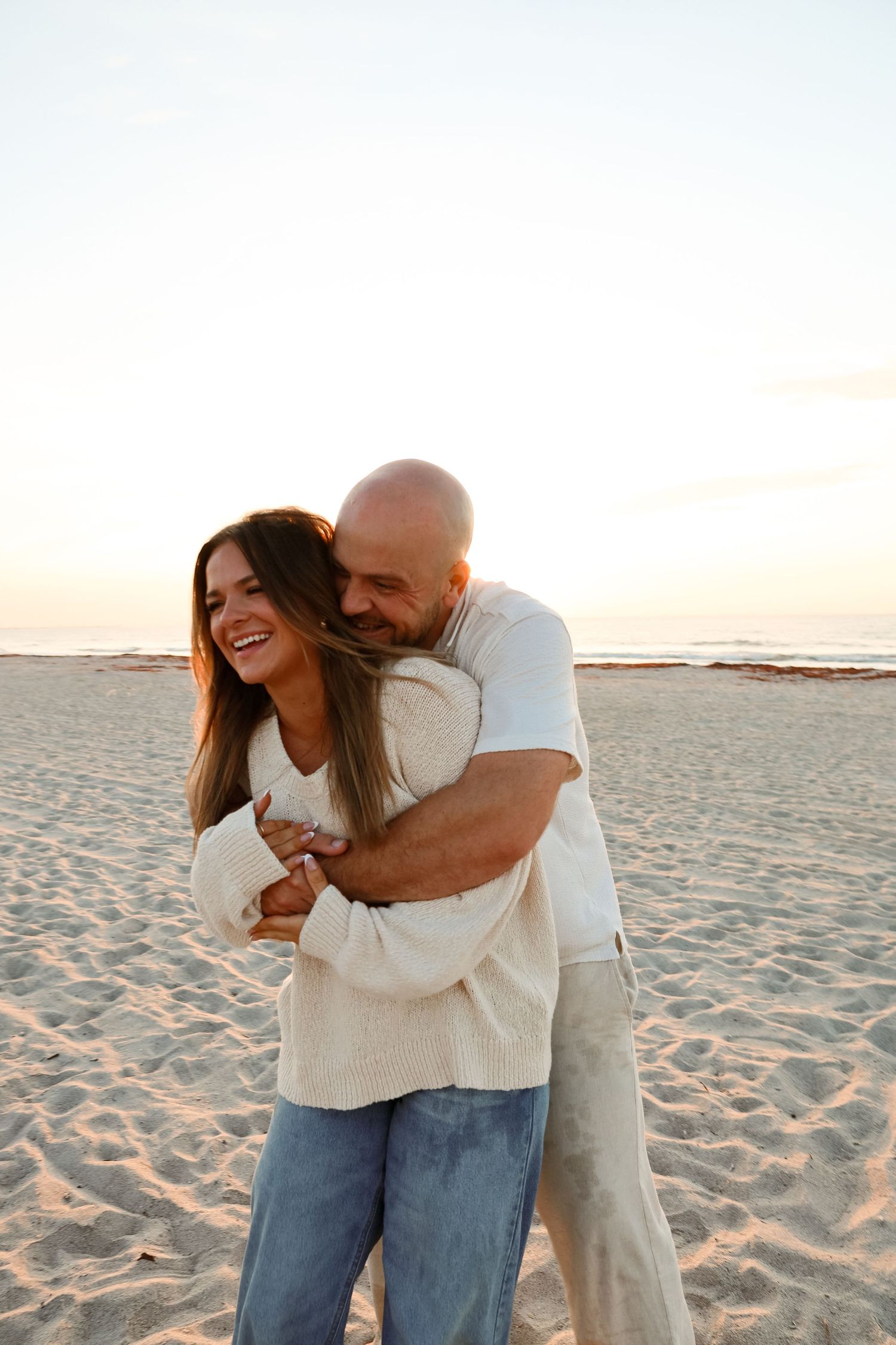 A happy couple embraces on a sandy beach at sunset, the woman laughing joyfully in a cozy sweater and jeans.