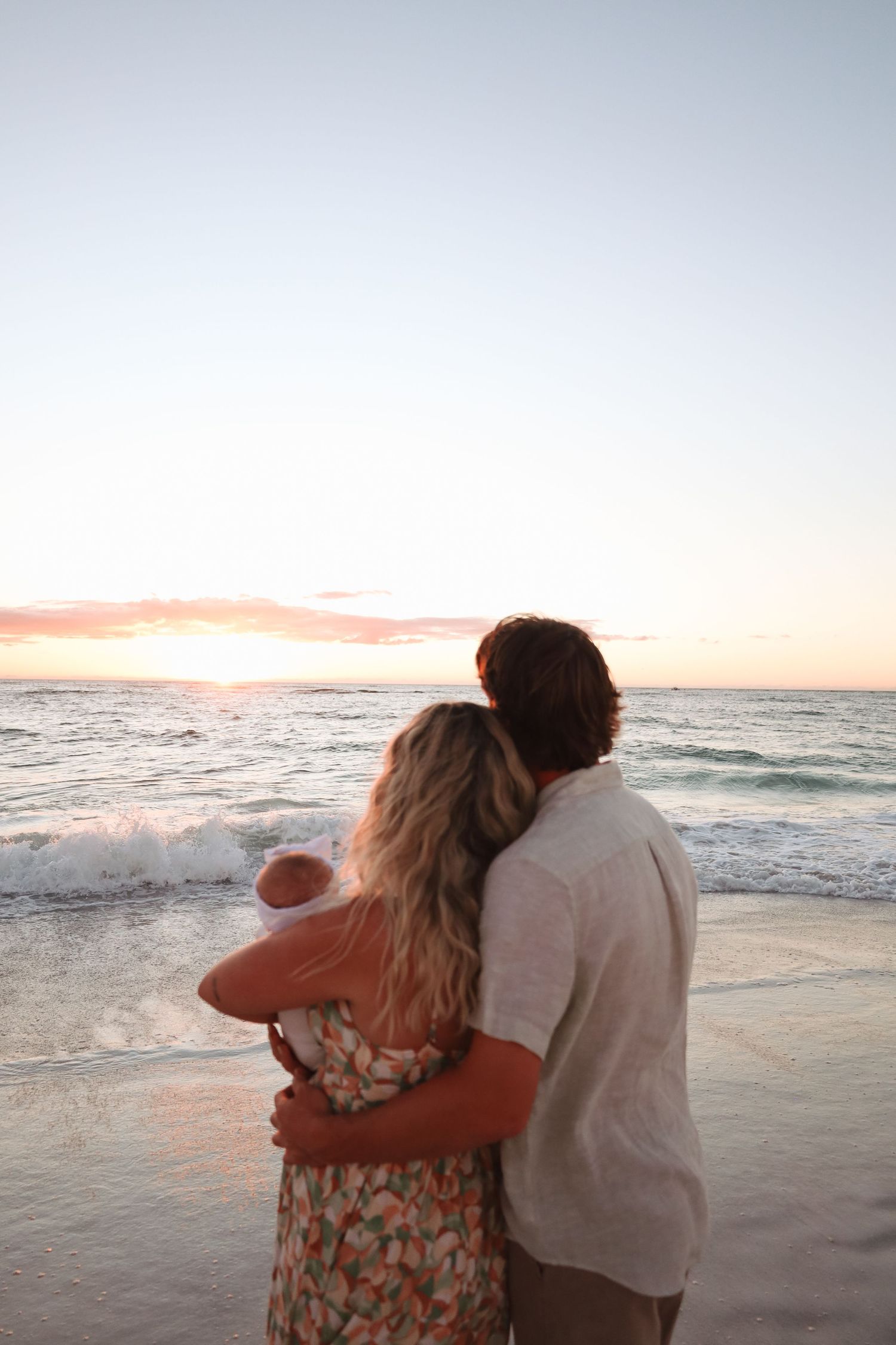 Couple embraces while watching sunset on a beach with waves crashing in the background.