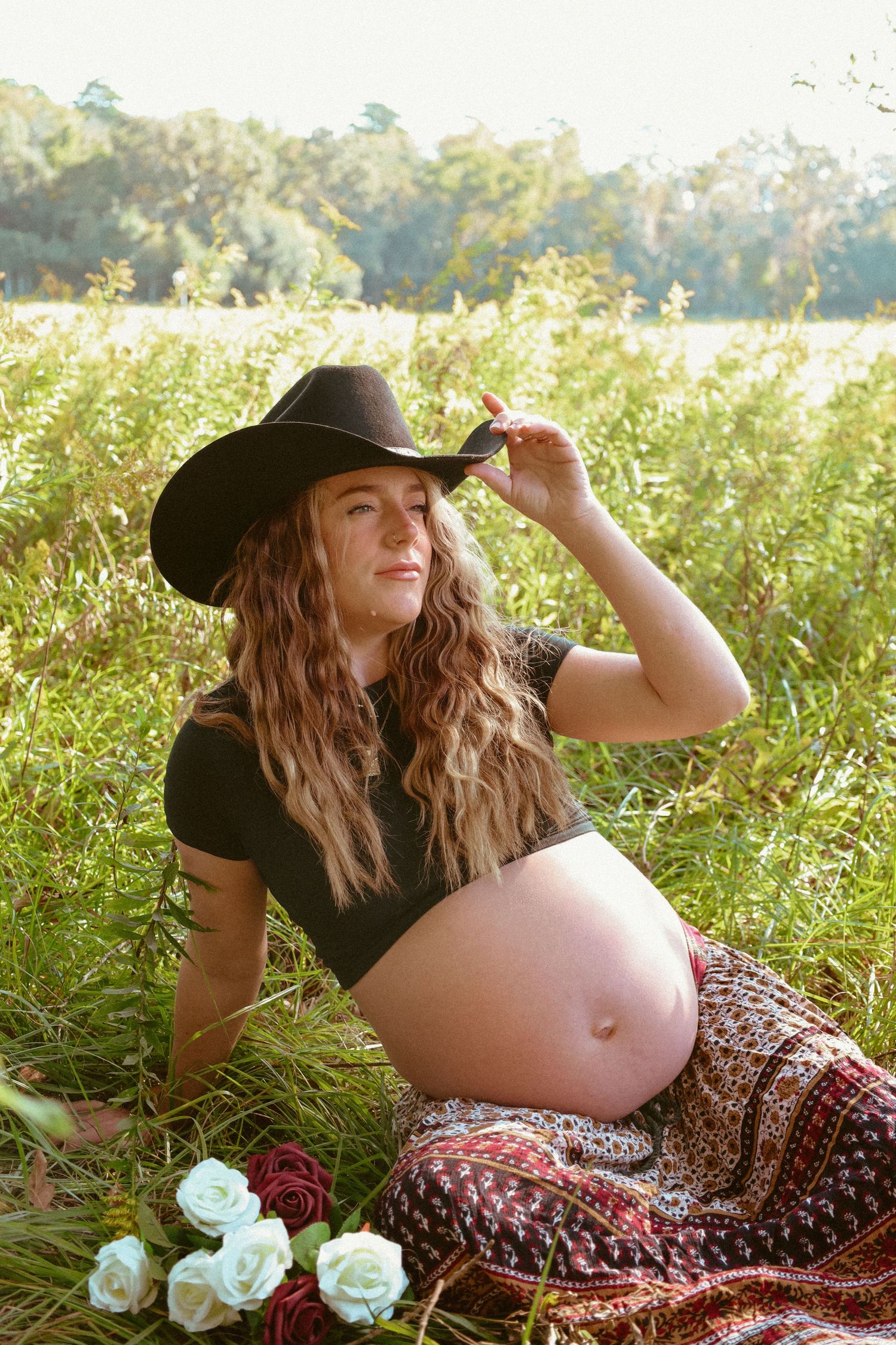 An expectant mother reclines in a sunlit field wearing a black hat and floral skirt.