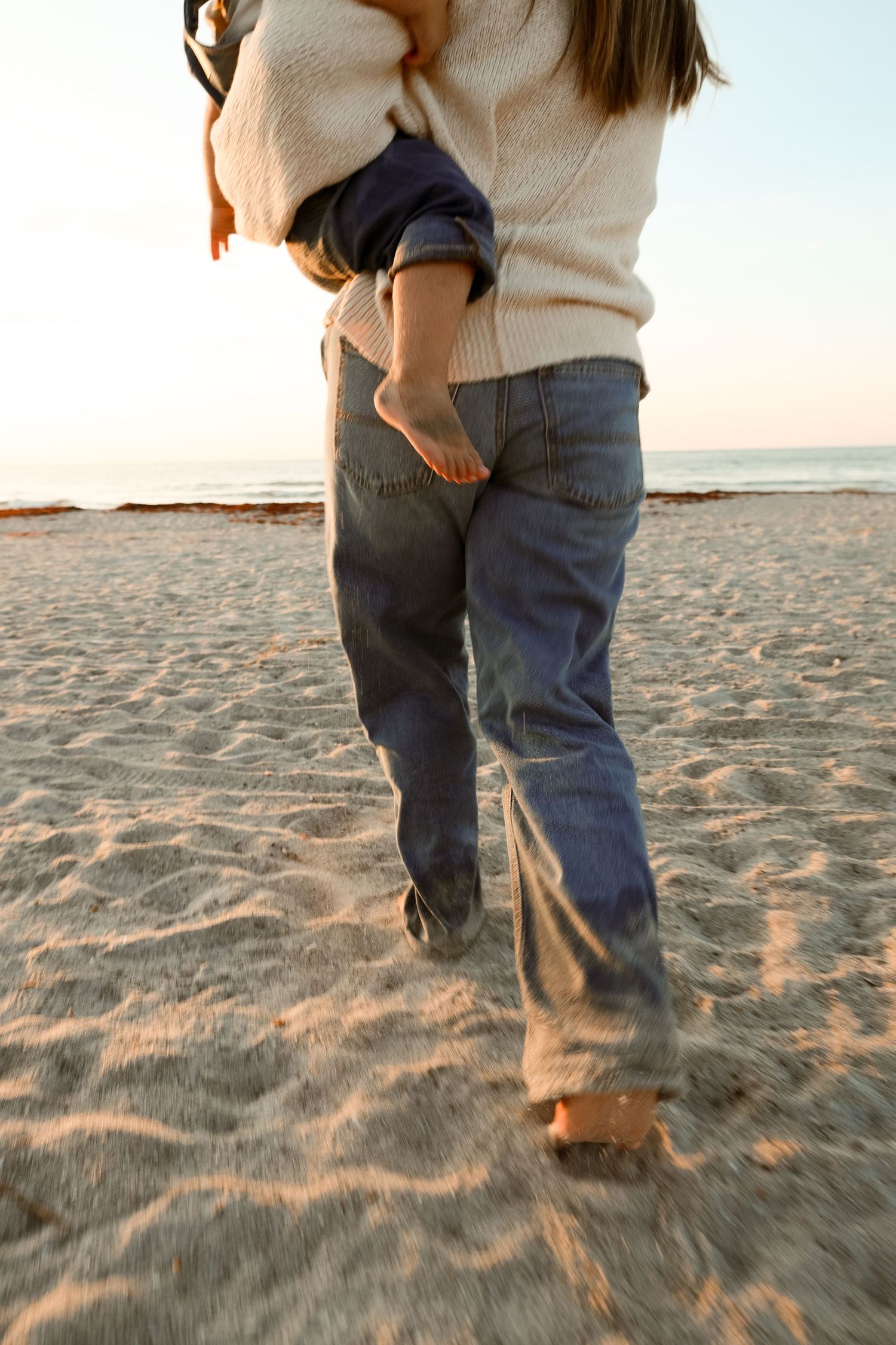 A parent carries a barefoot child on their hip while walking on a sandy beach at sunset, both wearing jeans.