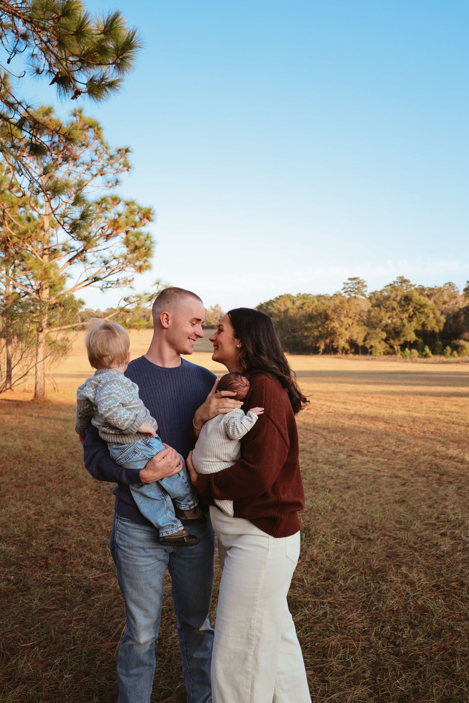 A family shares a tender moment together in a field at sunset with pine trees in the background.