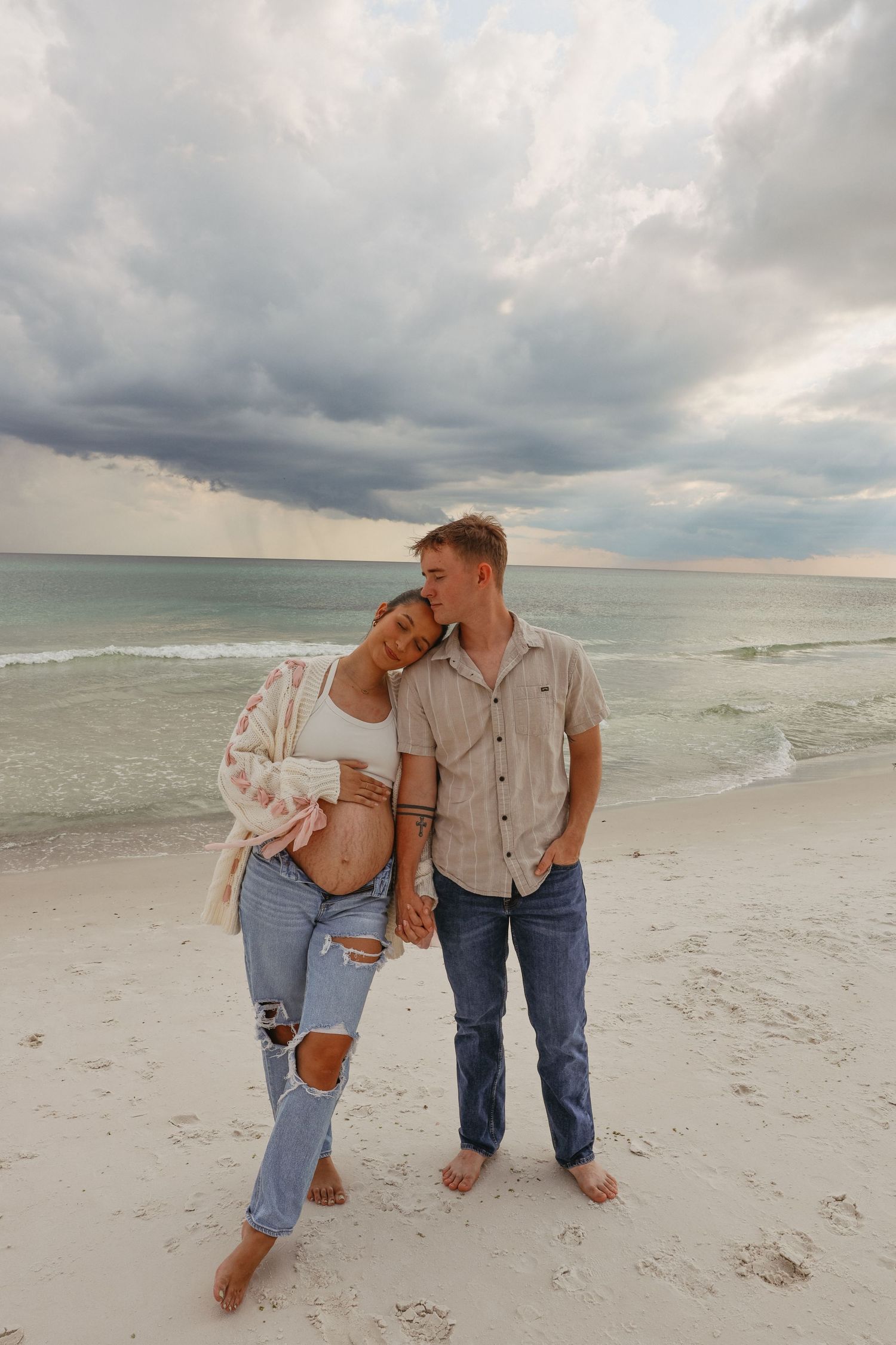 A couple stands together on a white sandy beach with dramatic storm clouds overhead and ocean waves in the background.