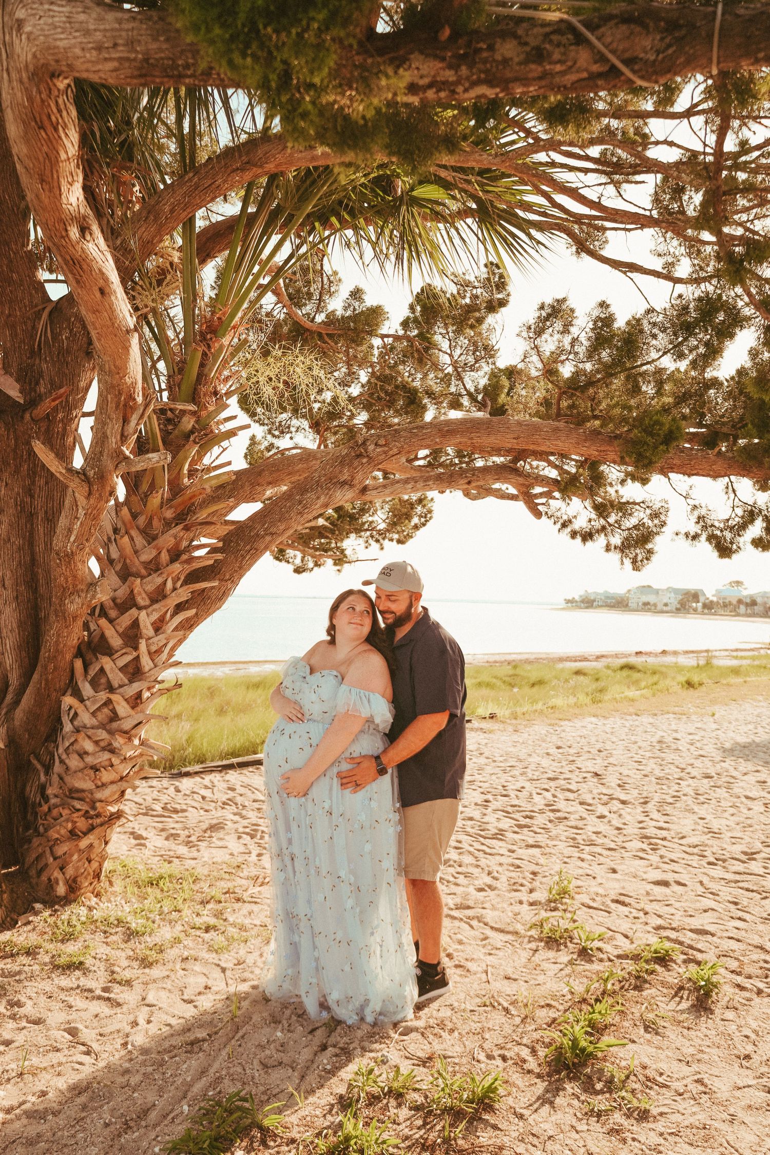 A maternity photo of an expectant couple under a palm tree on a beach at sunset.