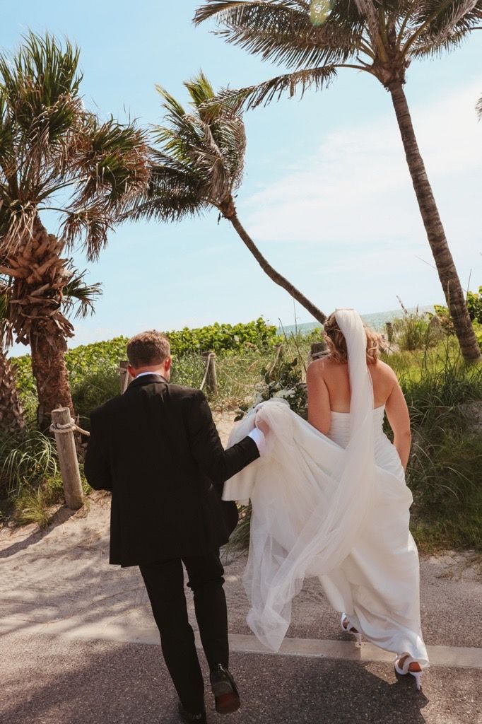 Bride and groom walk together on a sandy beach path lined with palm trees and tropical foliage on their wedding day.