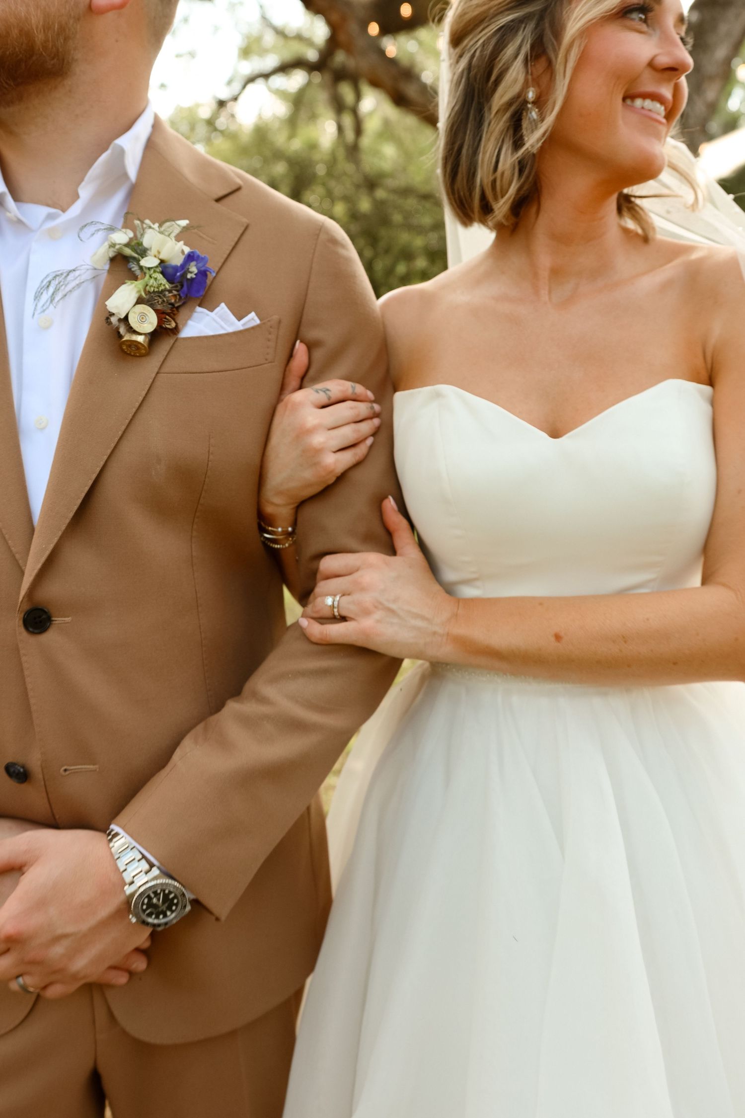 Bride in strapless white tulle gown holds groom's arm in tan suit with floral boutonniere at outdoor wedding.