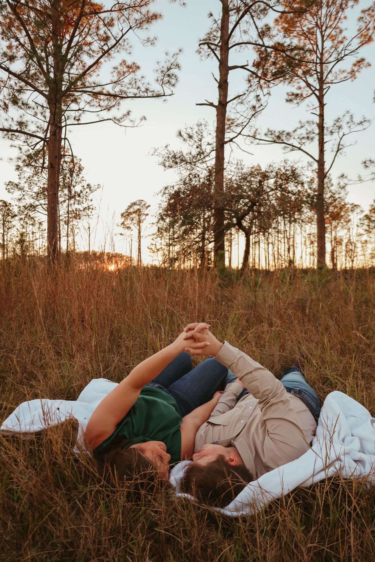 A couple lies together on a blanket in tall grass at sunset with tall pine trees silhouetted in the background.
