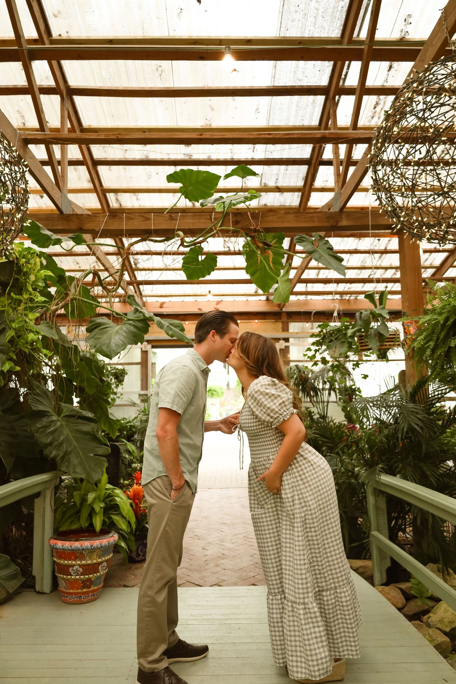A couple shares a romantic kiss on a wooden walkway inside a lush greenhouse with tropical plants.