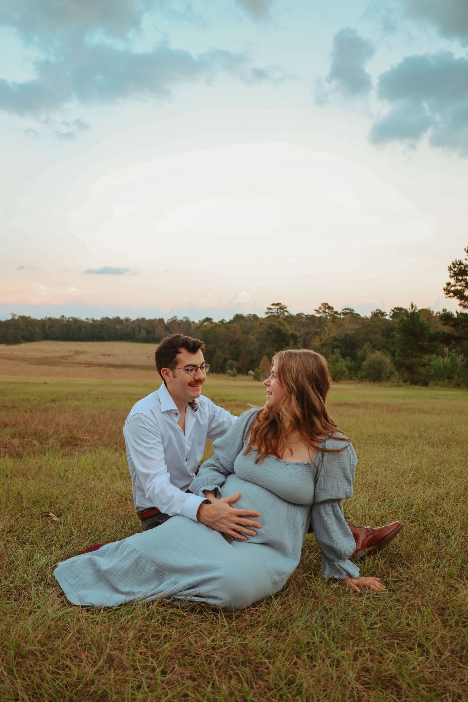 A couple in casual attire lies together in a grassy field during golden hour.