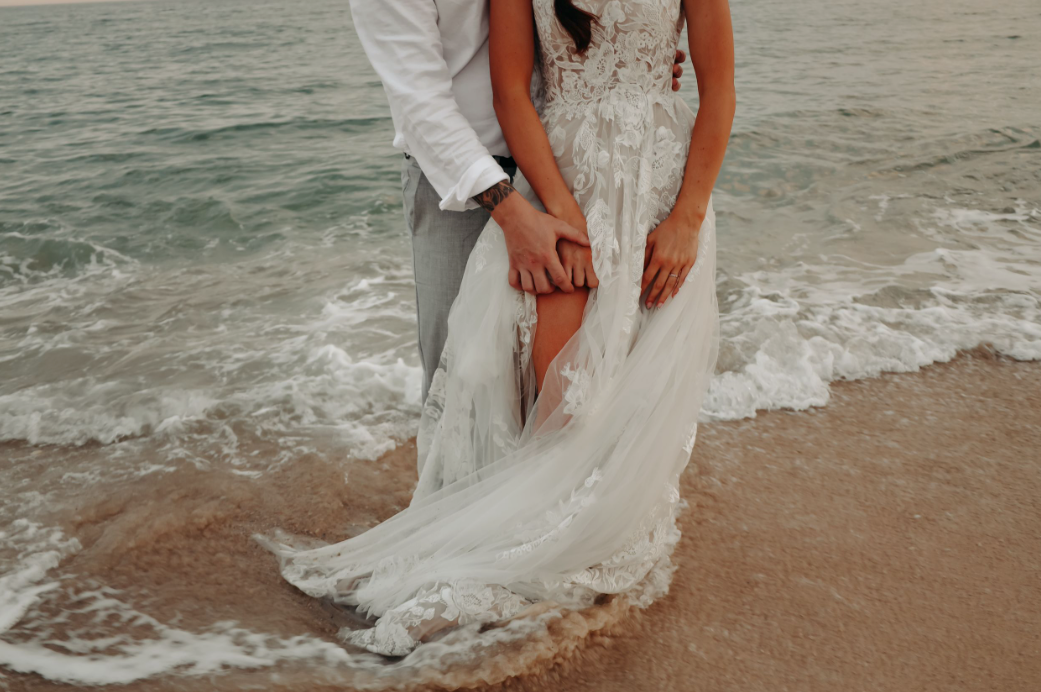 Ocean waves gently washing over white wedding dress on sandy beach.