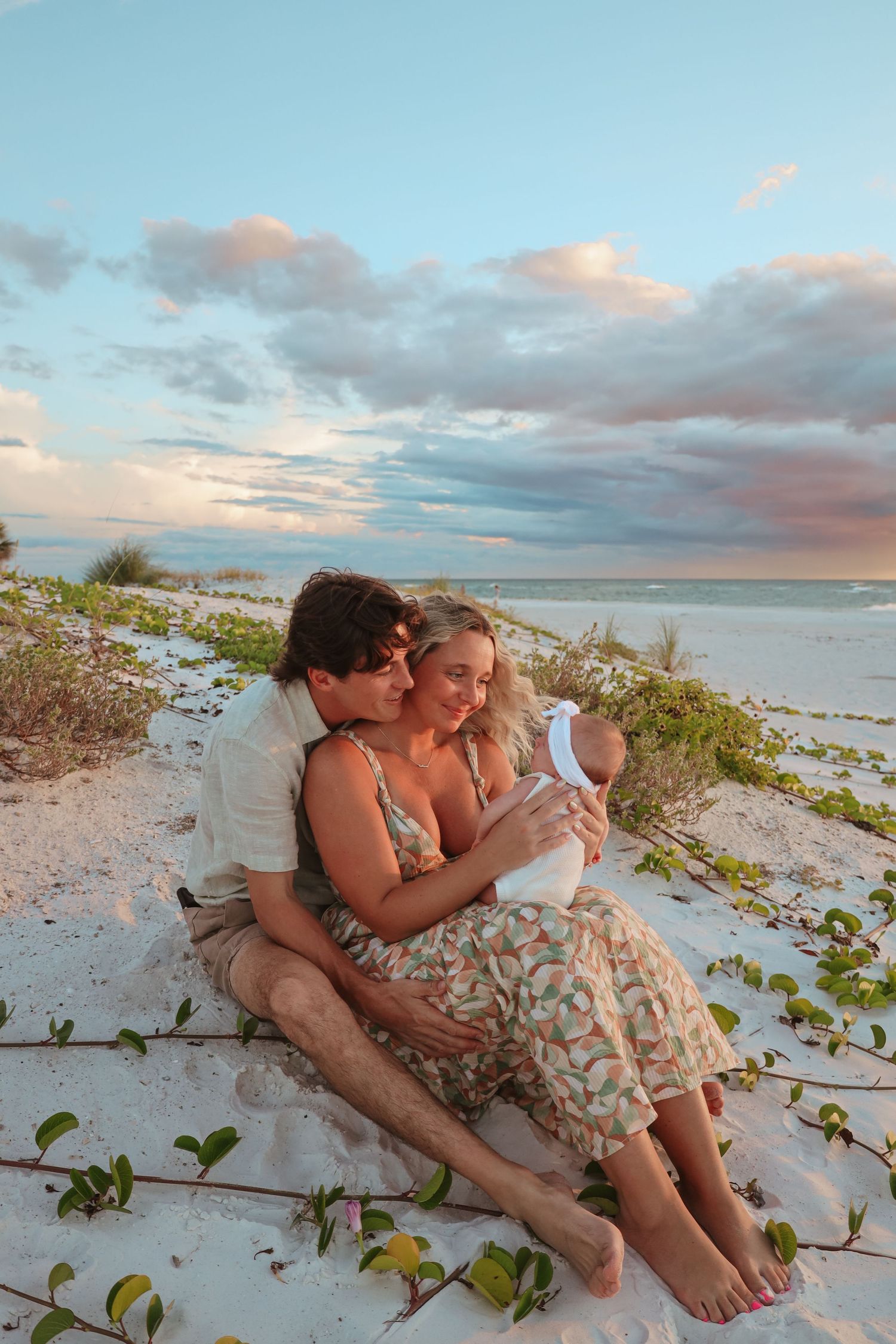 A couple embraces on a sandy beach at sunset with coastal vegetation in the foreground.