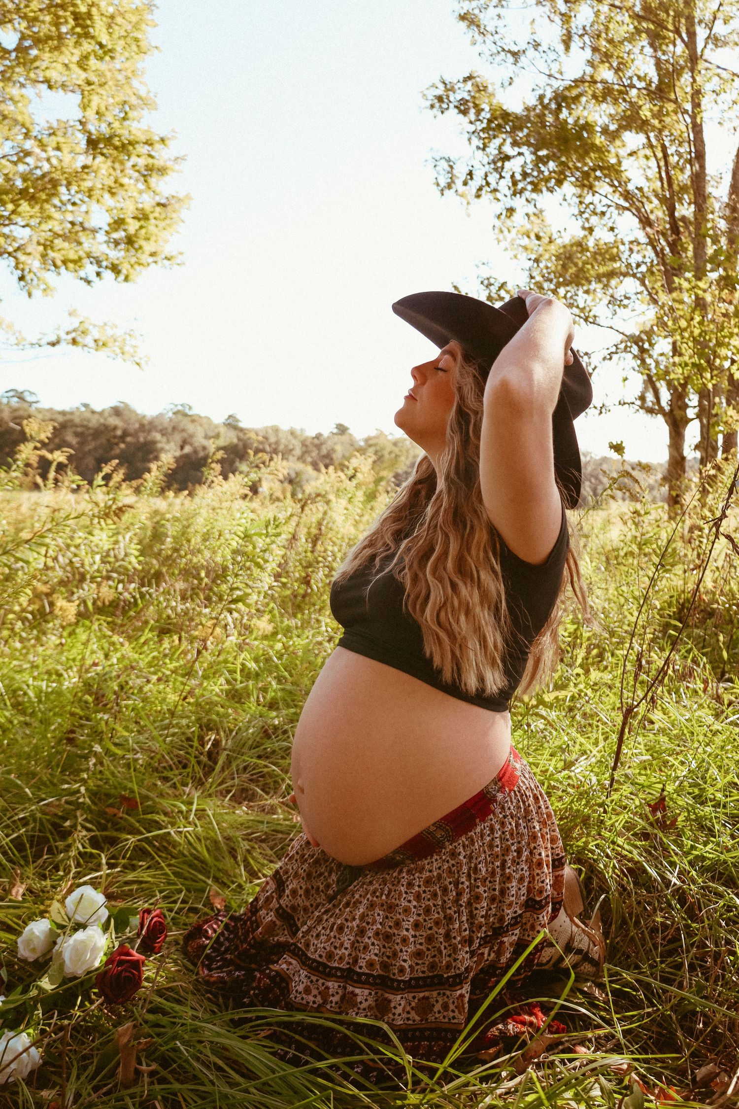 Pregnant woman in floral skirt kneels in sunlit meadow during golden hour maternity photoshoot.