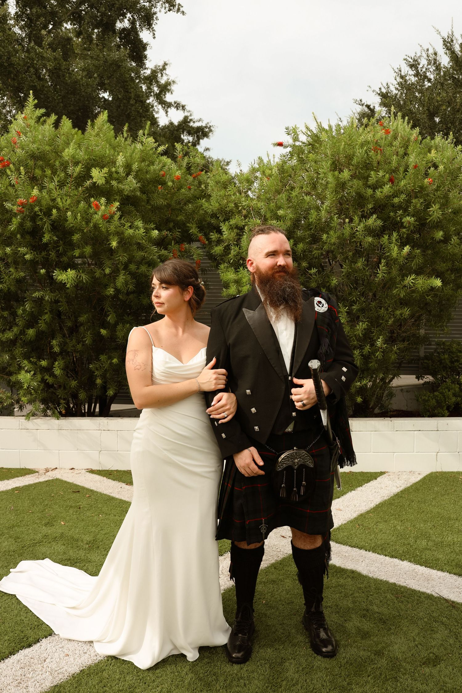 Bride in white gown and groom in black Scottish kilt with sporran pose together outdoors at wedding.