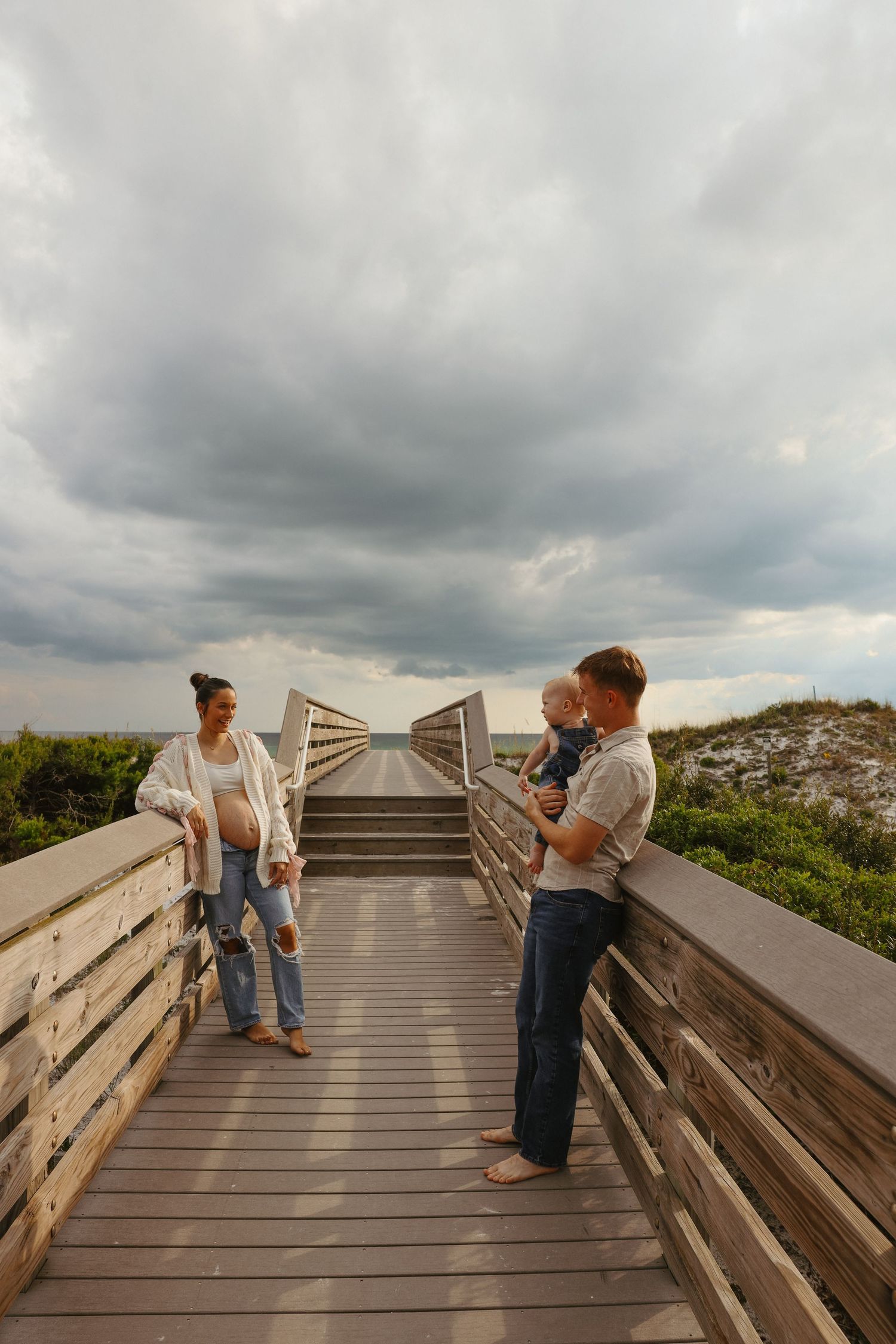 People gather on a wooden boardwalk overlooking a beach under dramatic cloudy skies.