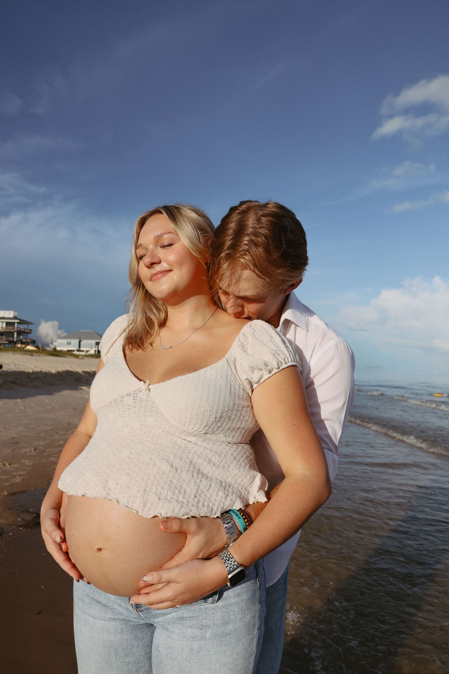 Pregnant couple embracing on a sunny beach, woman in white crop top and jeans with baby bump exposed.