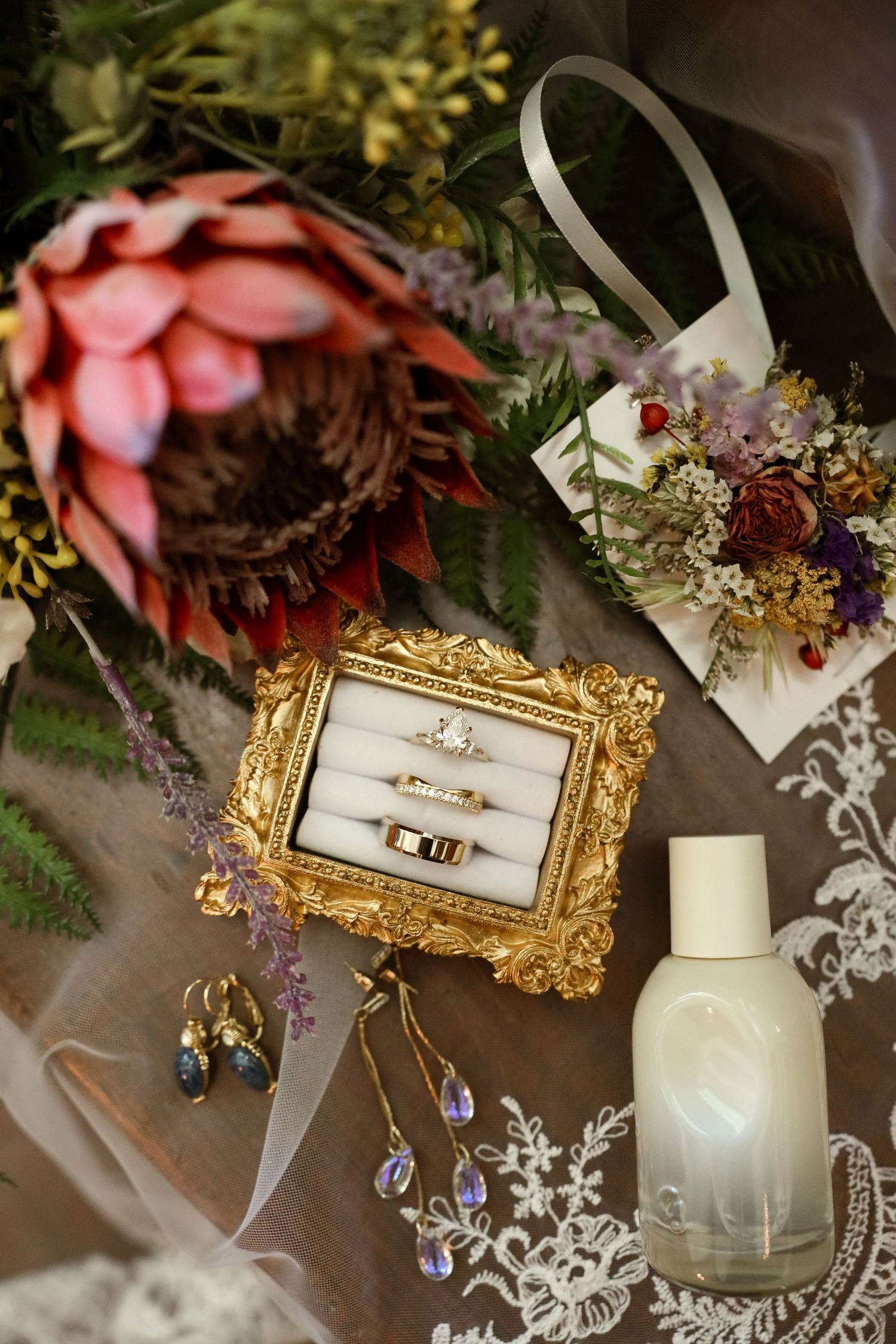 Gold ornate frame displaying wedding rings beside floral bouquet, perfume bottle, and crystal earrings on lace fabric.