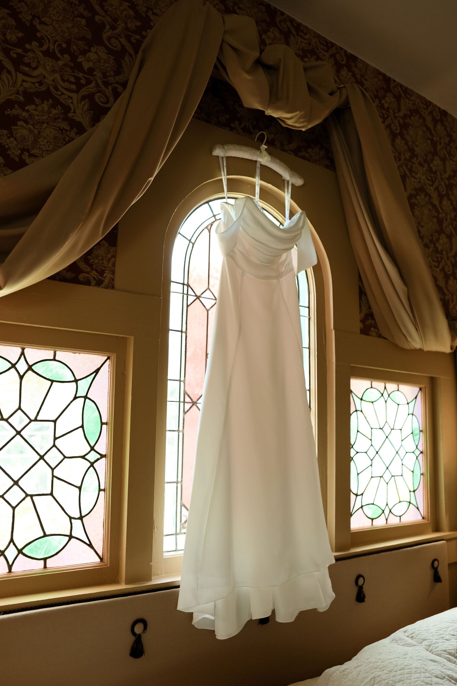 White strapless wedding dress hanging on hanger in front of arched window with stained glass panels.