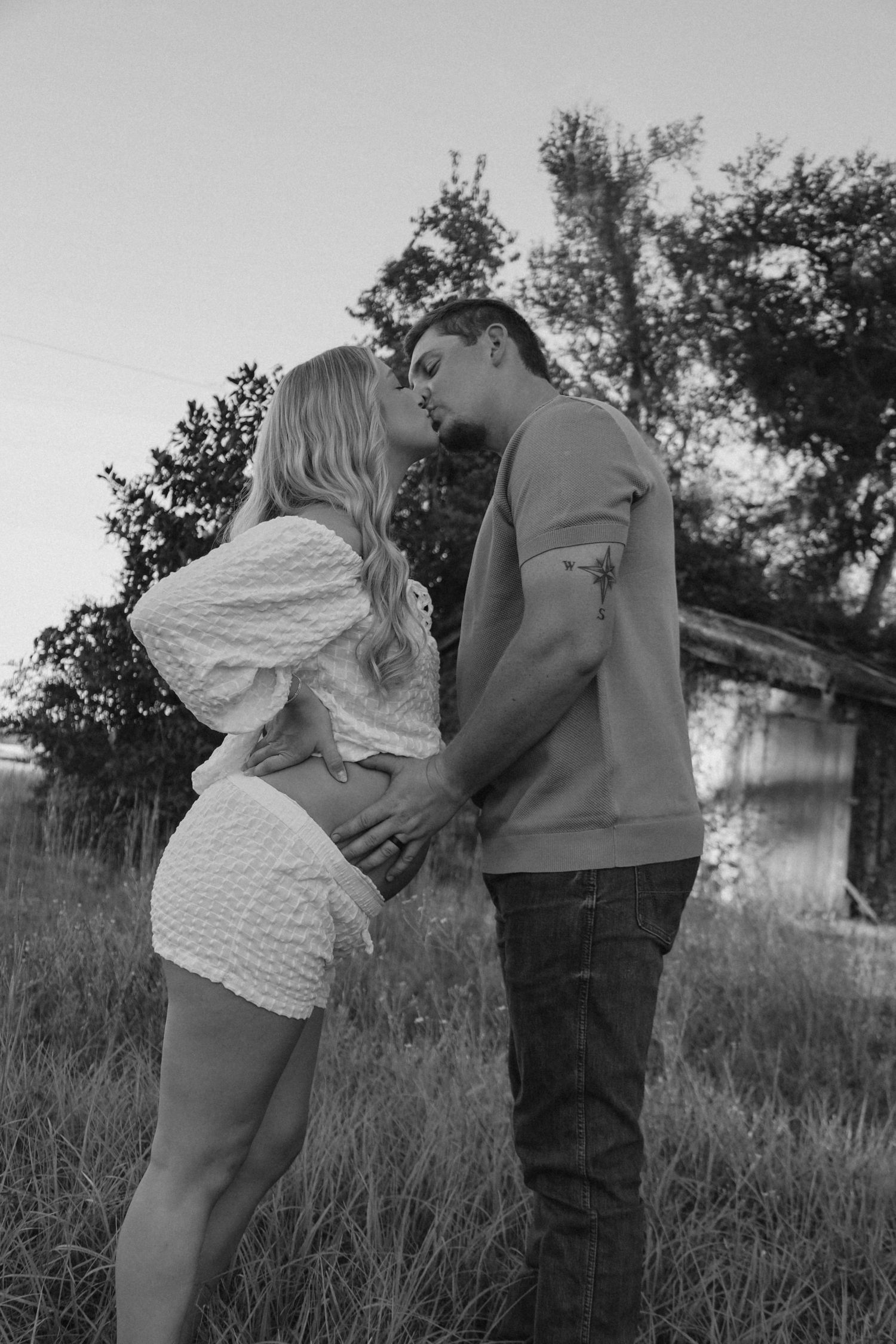 Black and white photo of a couple kissing in a grassy field at sunset, the man's hand resting on the woman's baby bump.