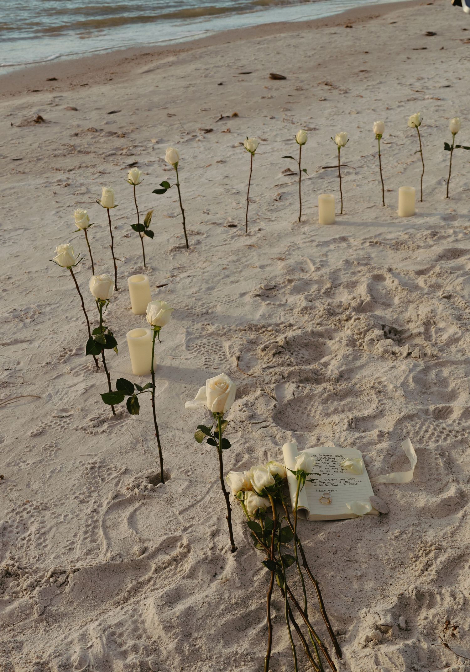 White roses and candles line up in a row along sandy beach at sunset creating a romantic memorial display.