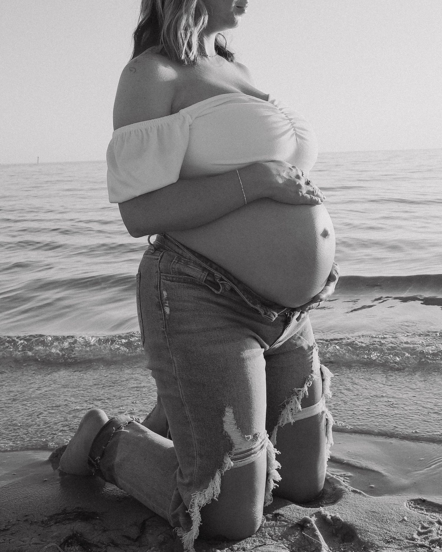 A person in ripped jeans sits by the ocean shoreline in a black and white artistic photograph.
