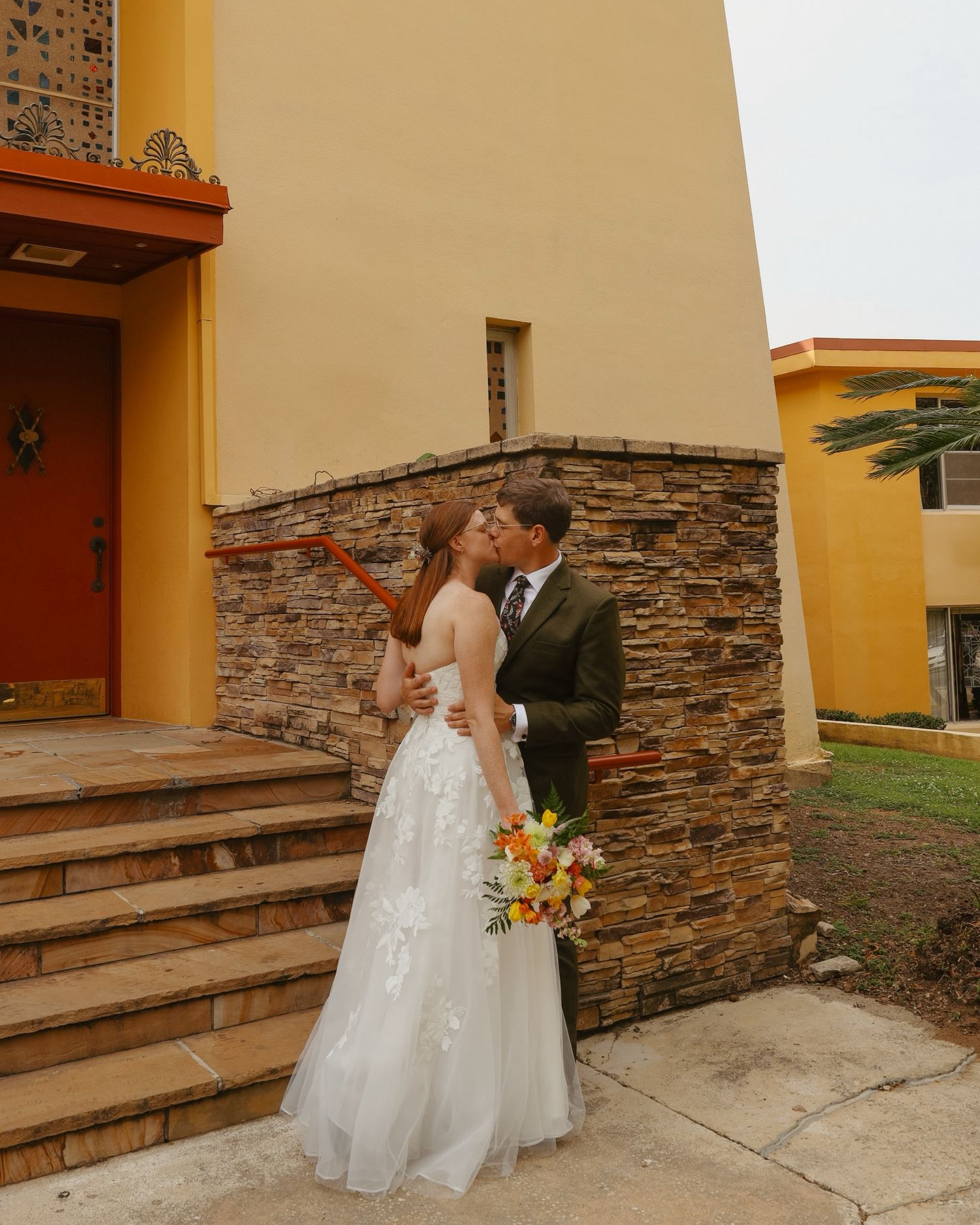 Wedding couple poses against stone wall exterior in formal attire.