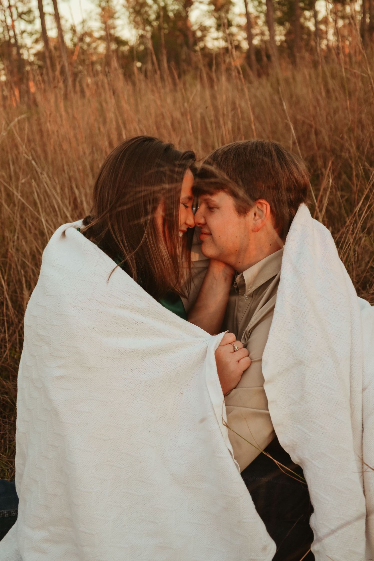 Romantic couple wrapped in white blanket share intimate moments during golden hour in tall grass field.