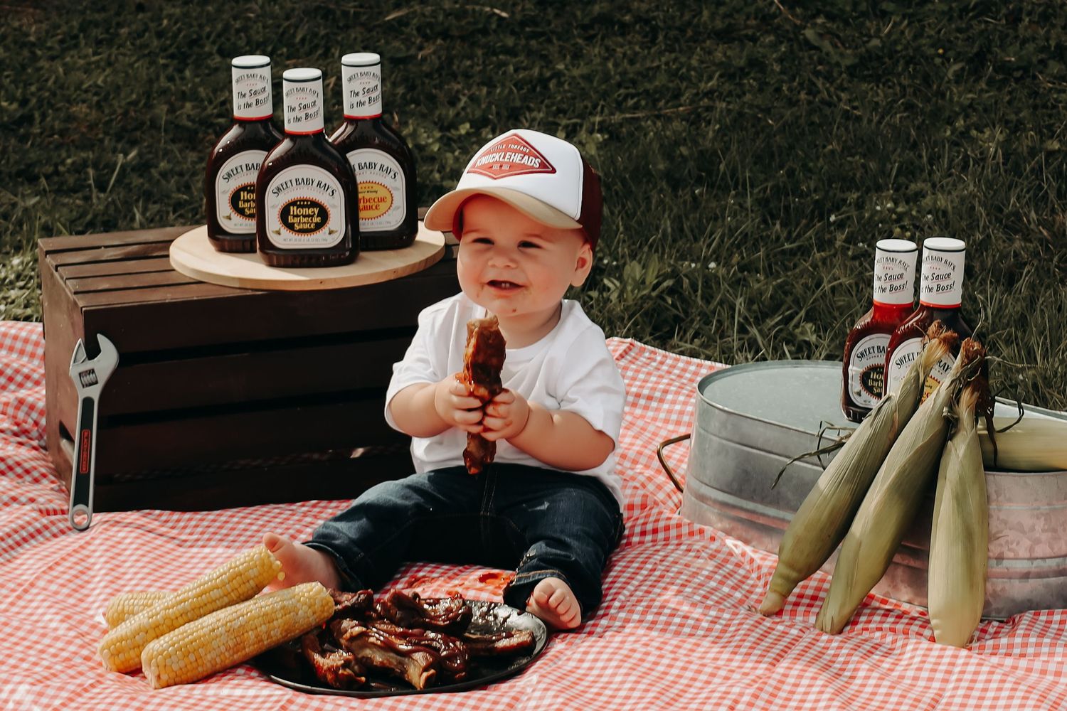 Picnic spread with fresh corn, meat, and checkered red tablecloth in an outdoor summer setting.