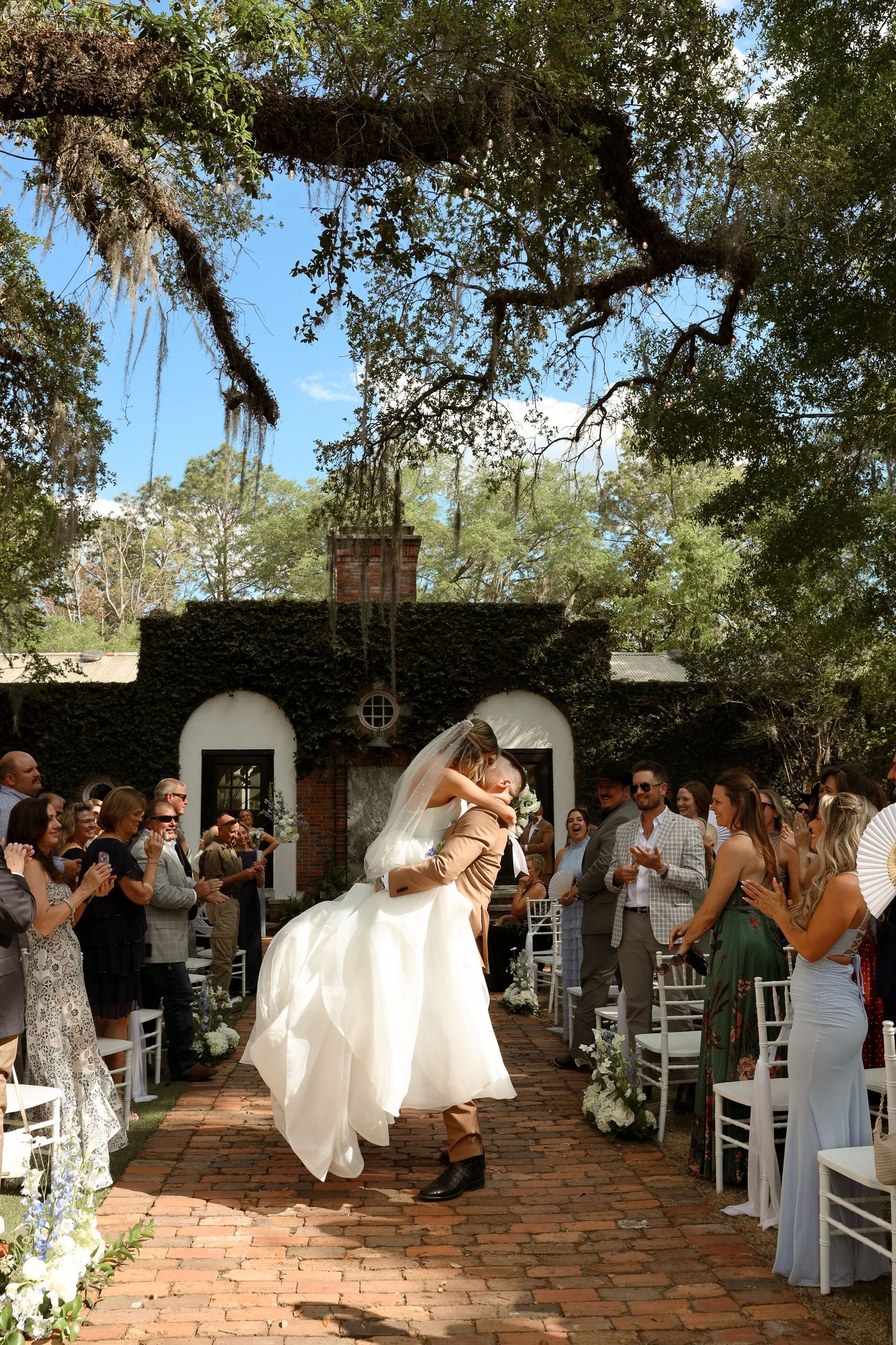 Groom lifts bride in white gown during outdoor wedding ceremony on brick aisle, surrounded by guests under oak trees.