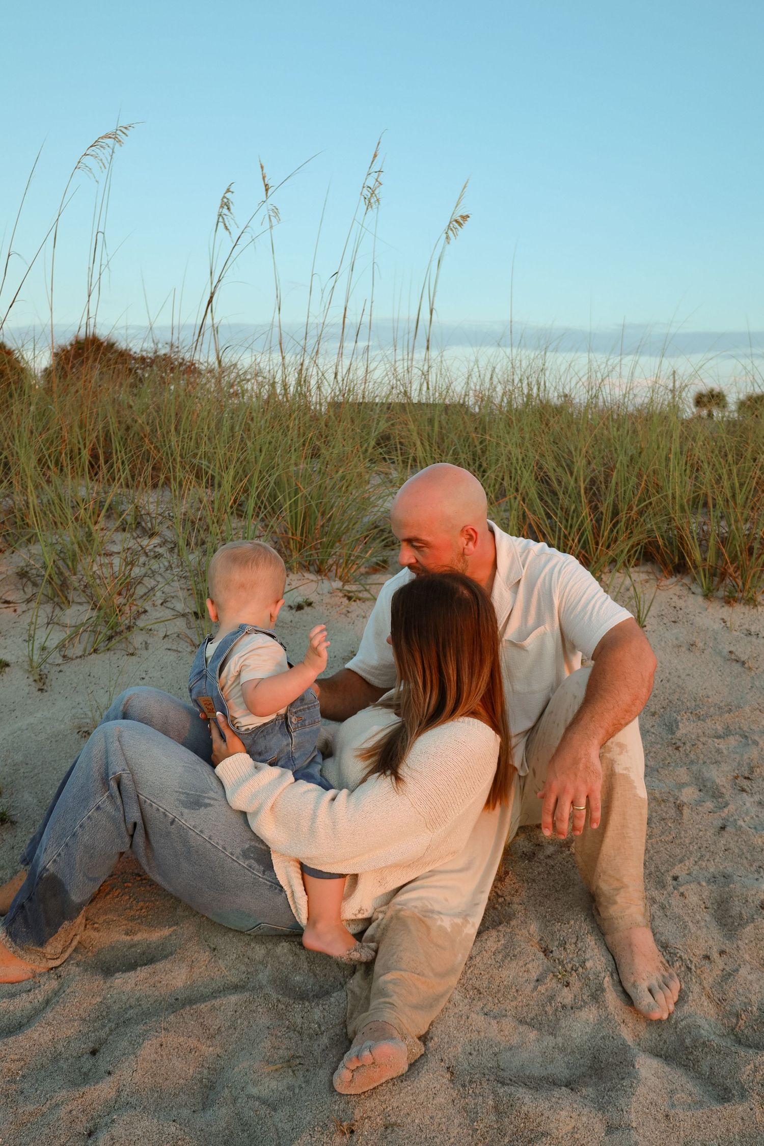 A family sits together on sandy beach dunes at sunset, with a bald father, mother, and baby in denim overalls enjoying the evening.